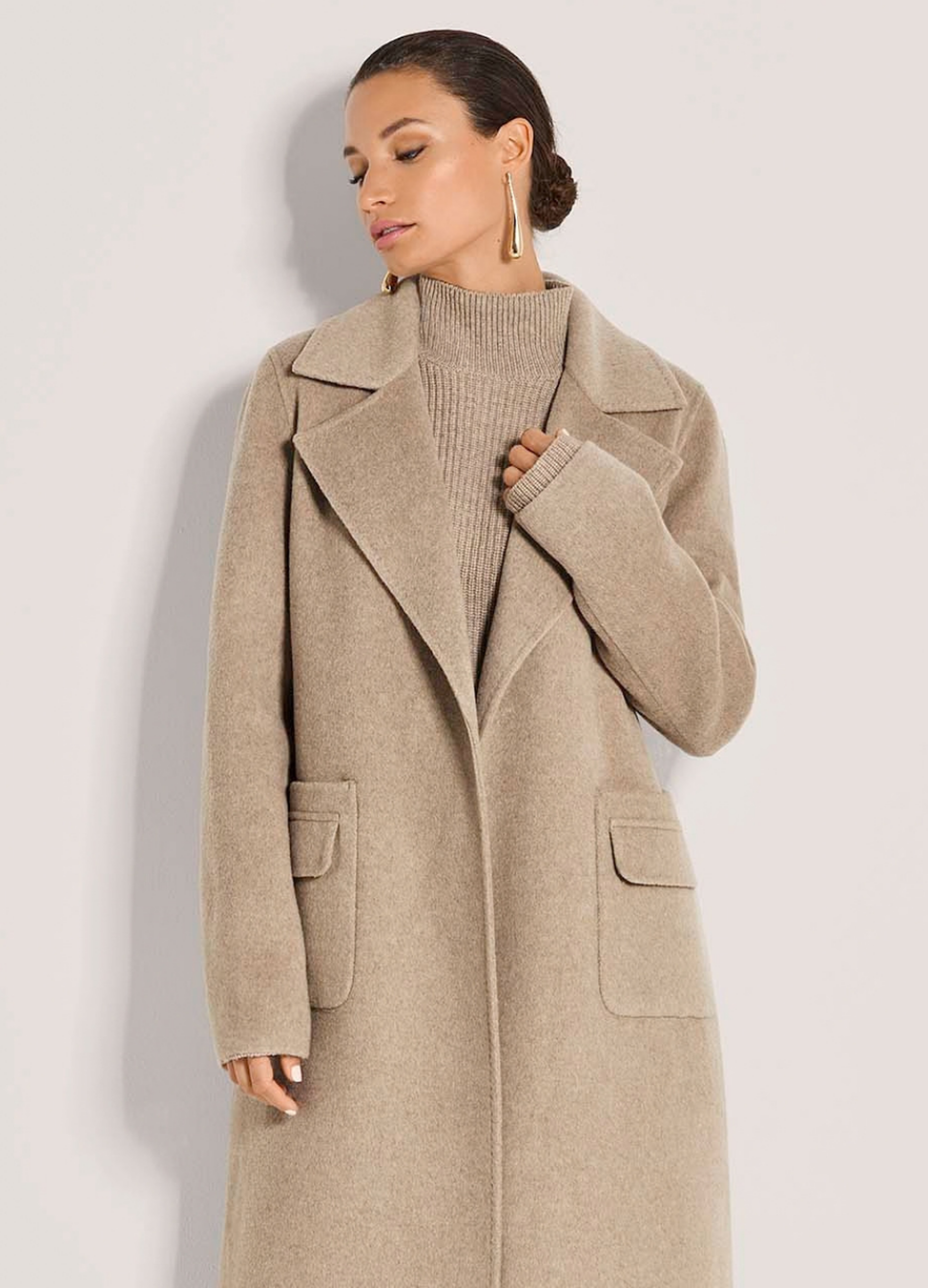 Frau im beigen Mantel mit breitem Revers und aufgesetzten Taschen über einem Strickpullover
