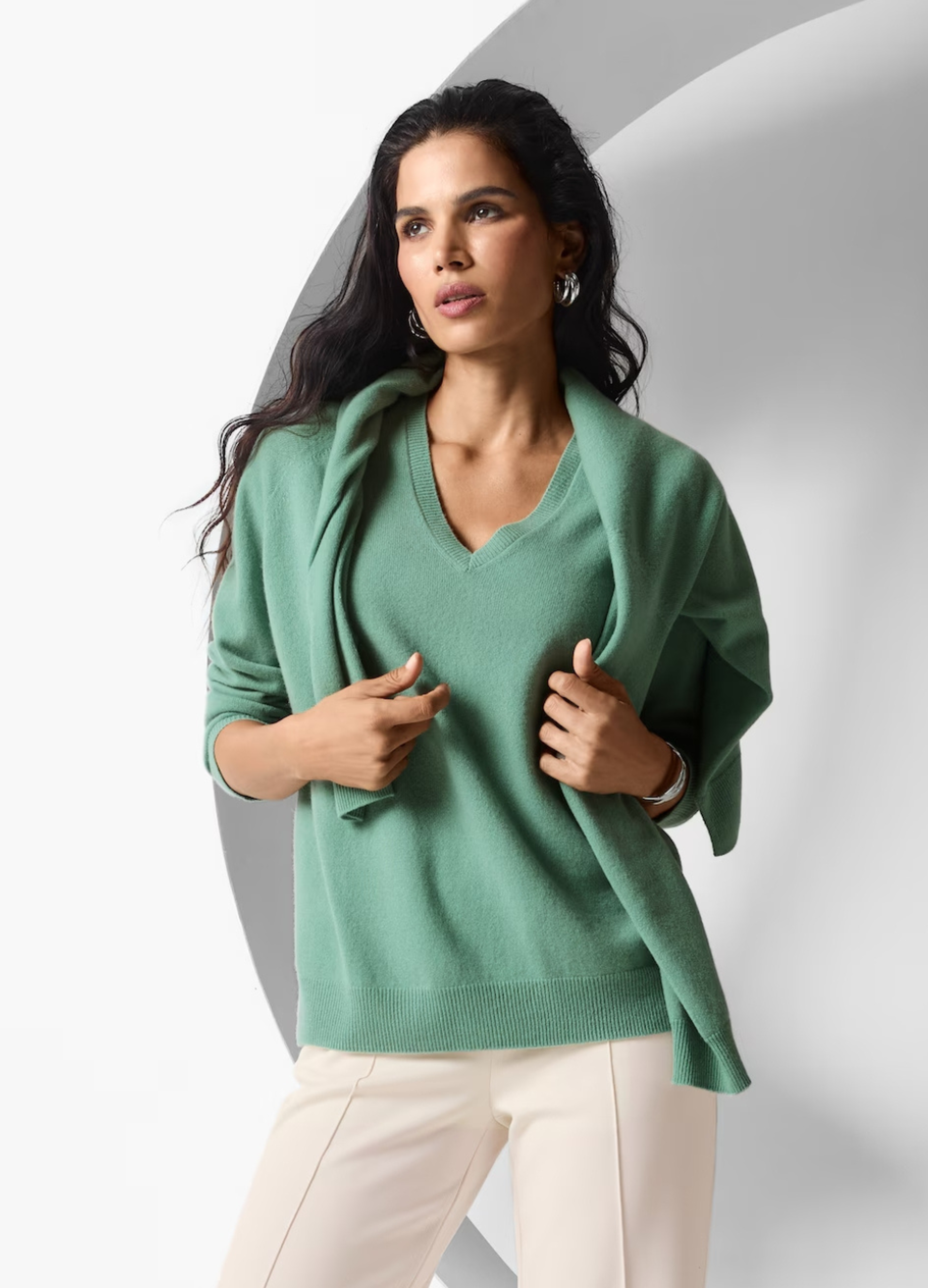 Eine Frau trägt einen hellsalbeigrünen Kaschmir-Pullover mit V-Ausschnitt. Passend dazu hat sie sich einen Cardigan in derselben Farbe über die Schultern gelegt.