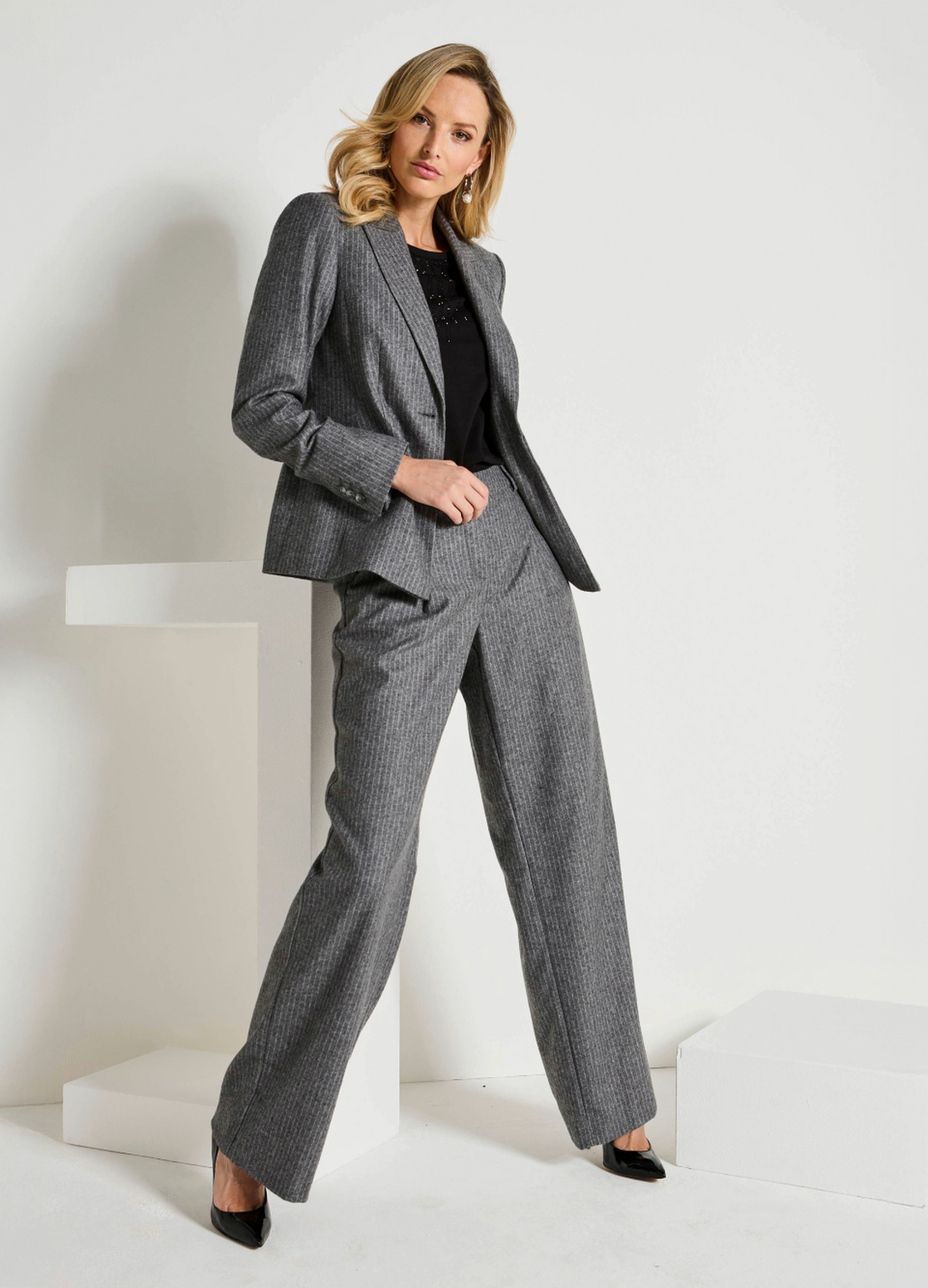 Die Frau trägt einen eleganten, grauen Hosenanzug mit weißen Nadelstreifen. Darunter trägt sie ein schwarzes Oberteil mit Glitzerdetails.