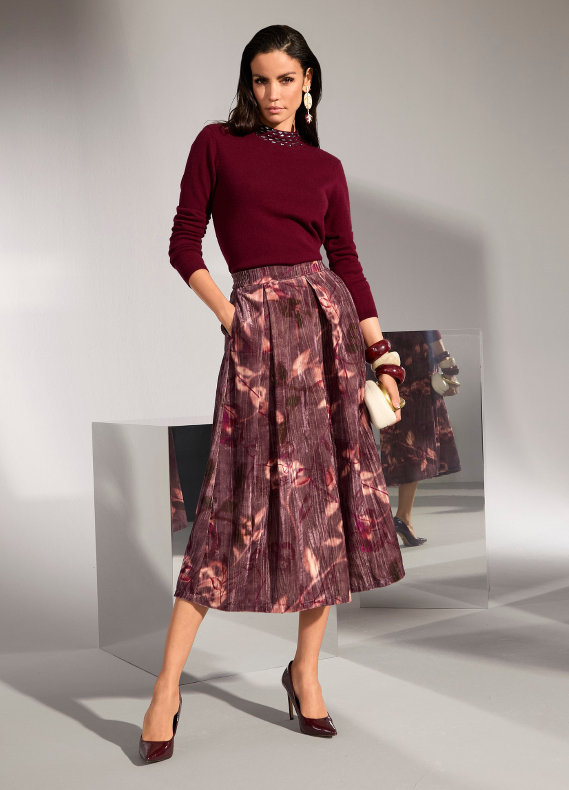 Eine Frau trägt einen bordeauxfarbenen Pullover mit glänzenden Details am Kragen. Dazu trägt sie einen Rock mit floralem Muster.