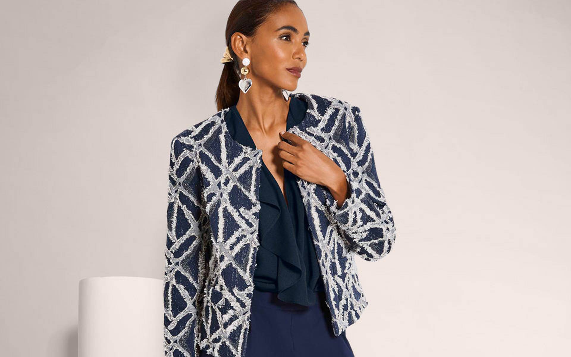 Donkerblauwe korte blazer met wit grafisch structuurpatroon, open gedragen over een blouse met volantdetail.