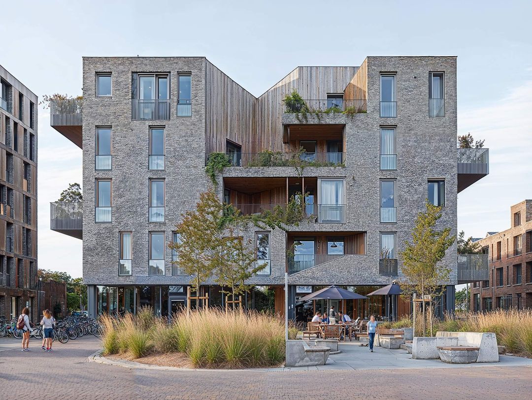 Modern multi-story building with brick and wood facade, featuring balconies, greenery, outdoor seating, and people walking nearby.