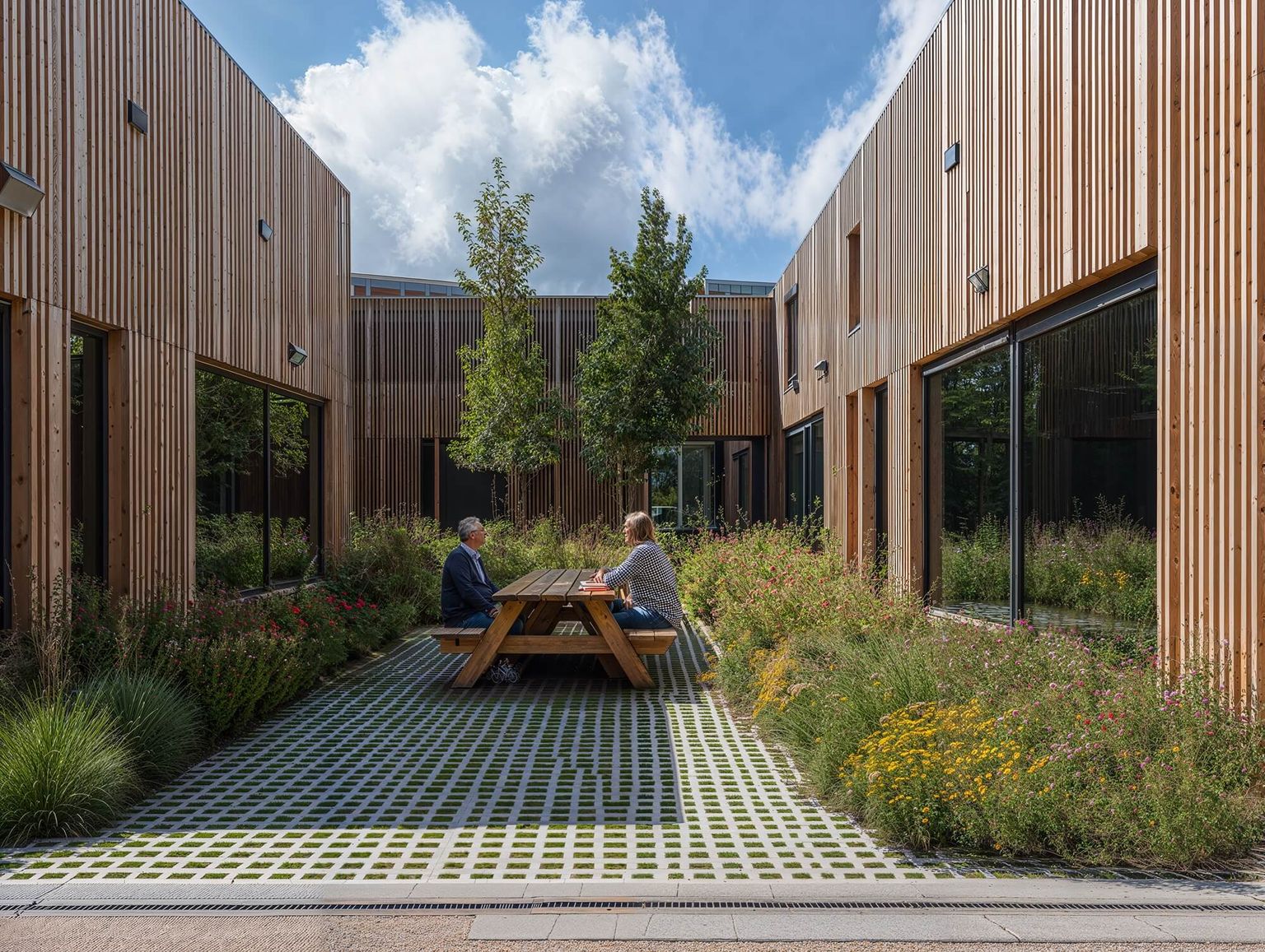 Two people sit at a picnic table in a modern courtyard with wooden buildings, surrounded by greenery and flowers under a partly cloudy sky.