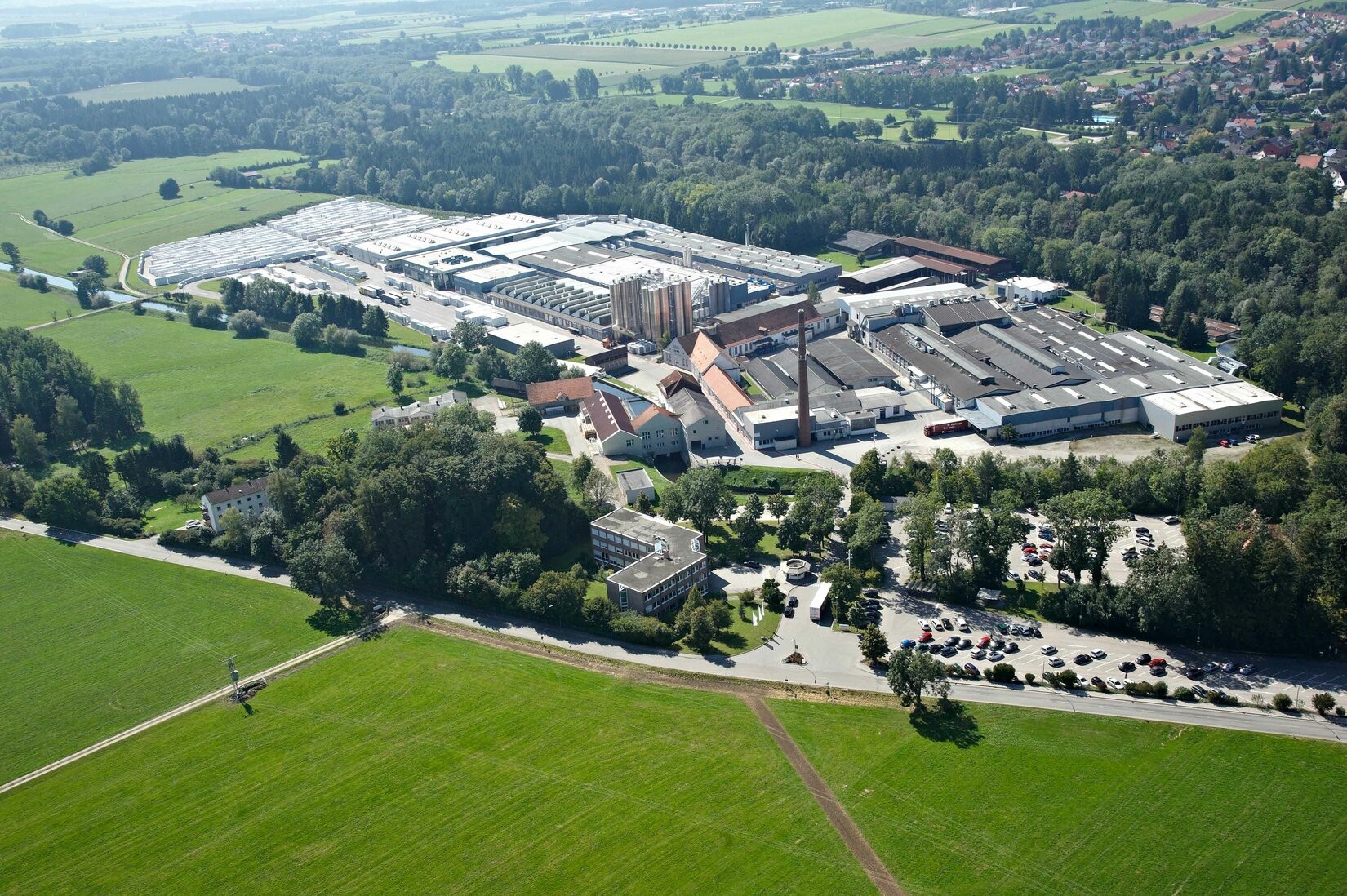 Una vista aérea de un gran complejo industrial rodeado de campos verdes y árboles, con un estacionamiento lleno de coches.