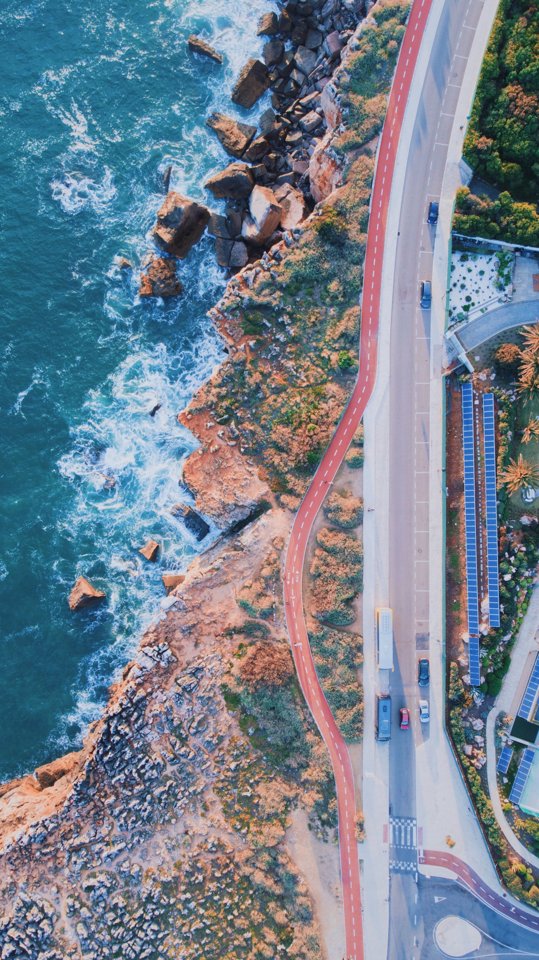 Vista aérea de una carretera costera con coches y camiones circulando por ella, rodeada de acantilados rocosos y el océano.