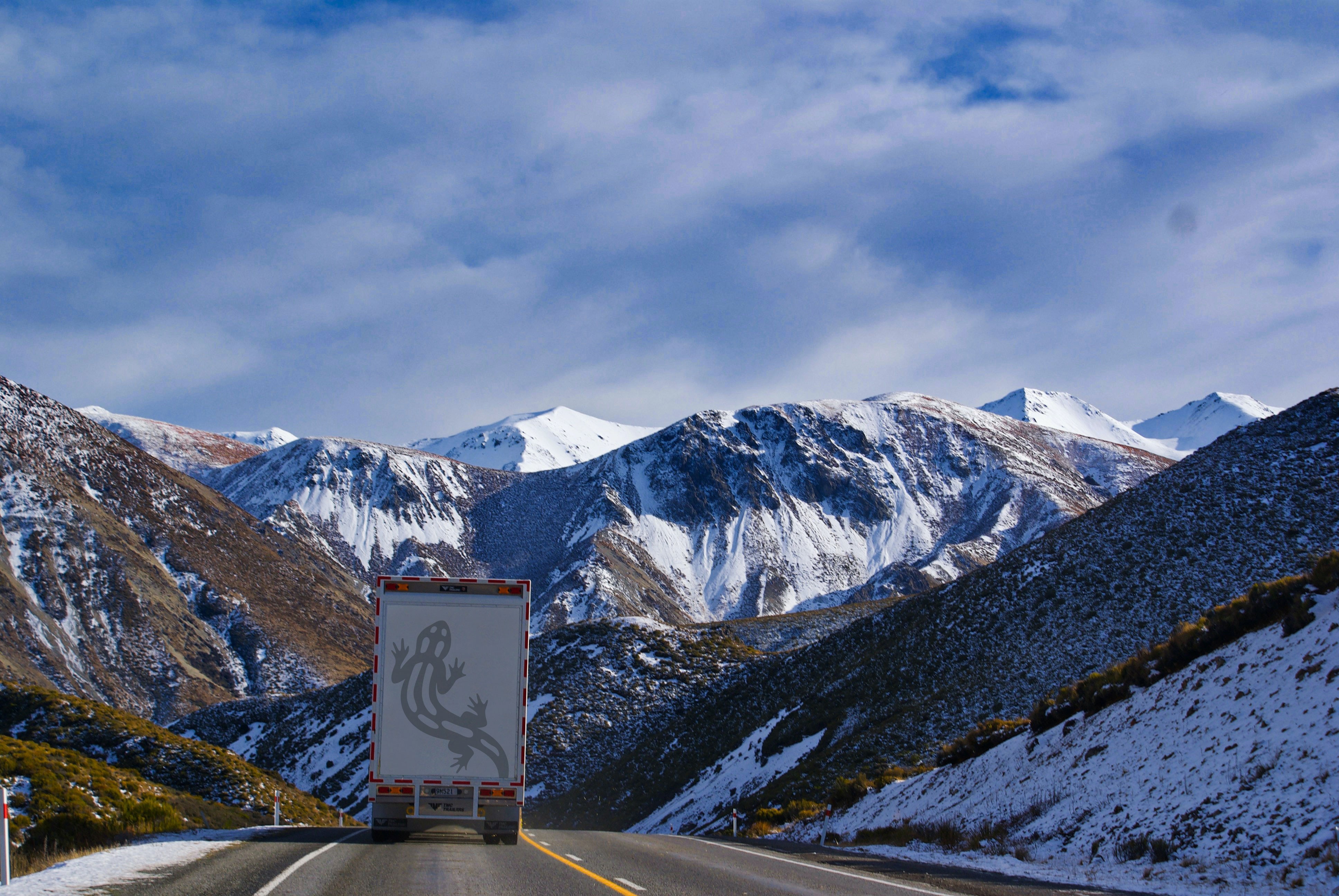 Un camión con un diseño de rana en la parte trasera conduce por una carretera de montaña nevada con un cielo azul y nubes blancas arriba.