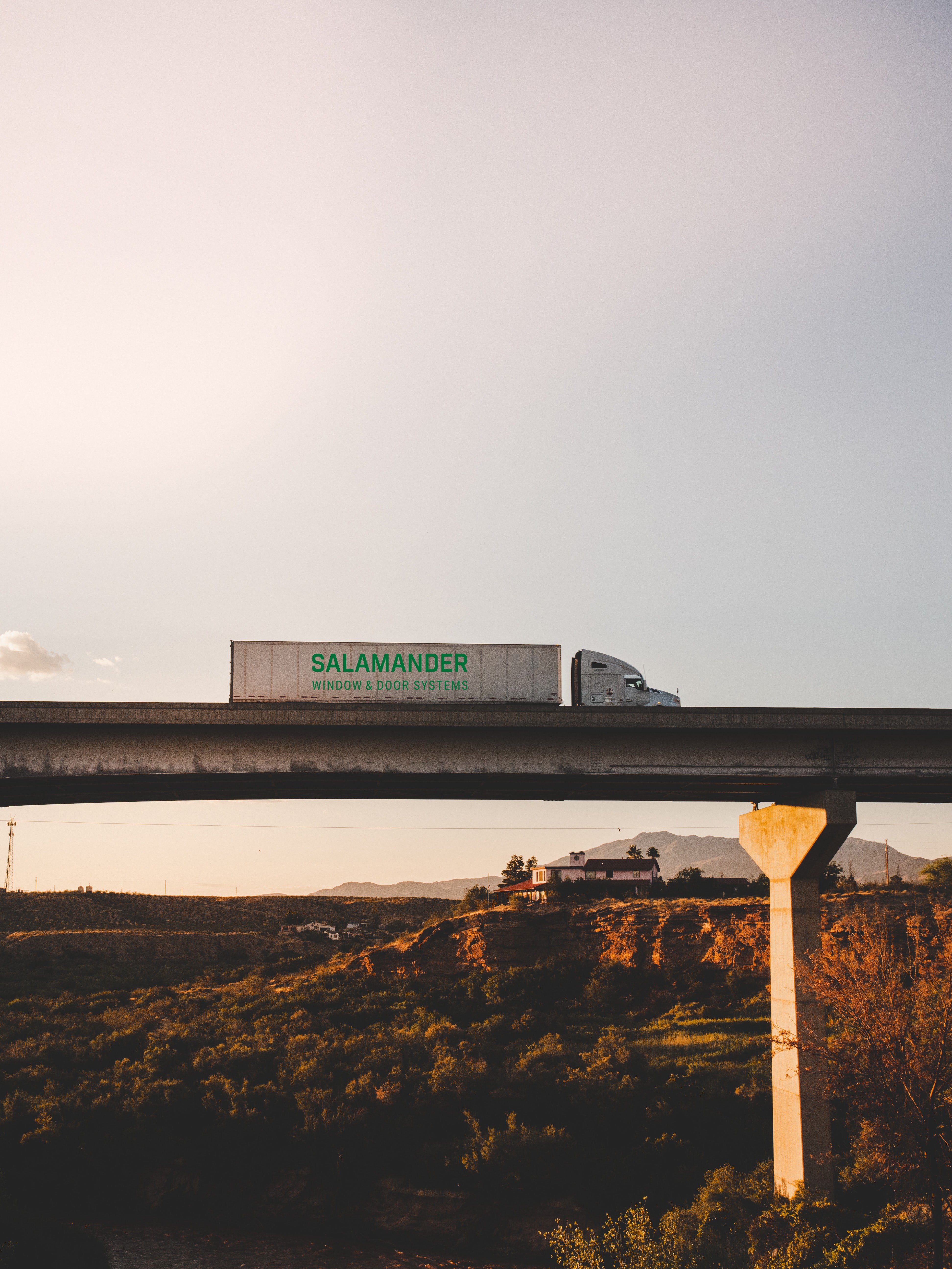 Un semi-camión blanco con un logotipo verde y las palabras "Salamander Window & Door Systems" en el costado está conduciendo sobre un puente que cruza un paisaje con montañas y casas.