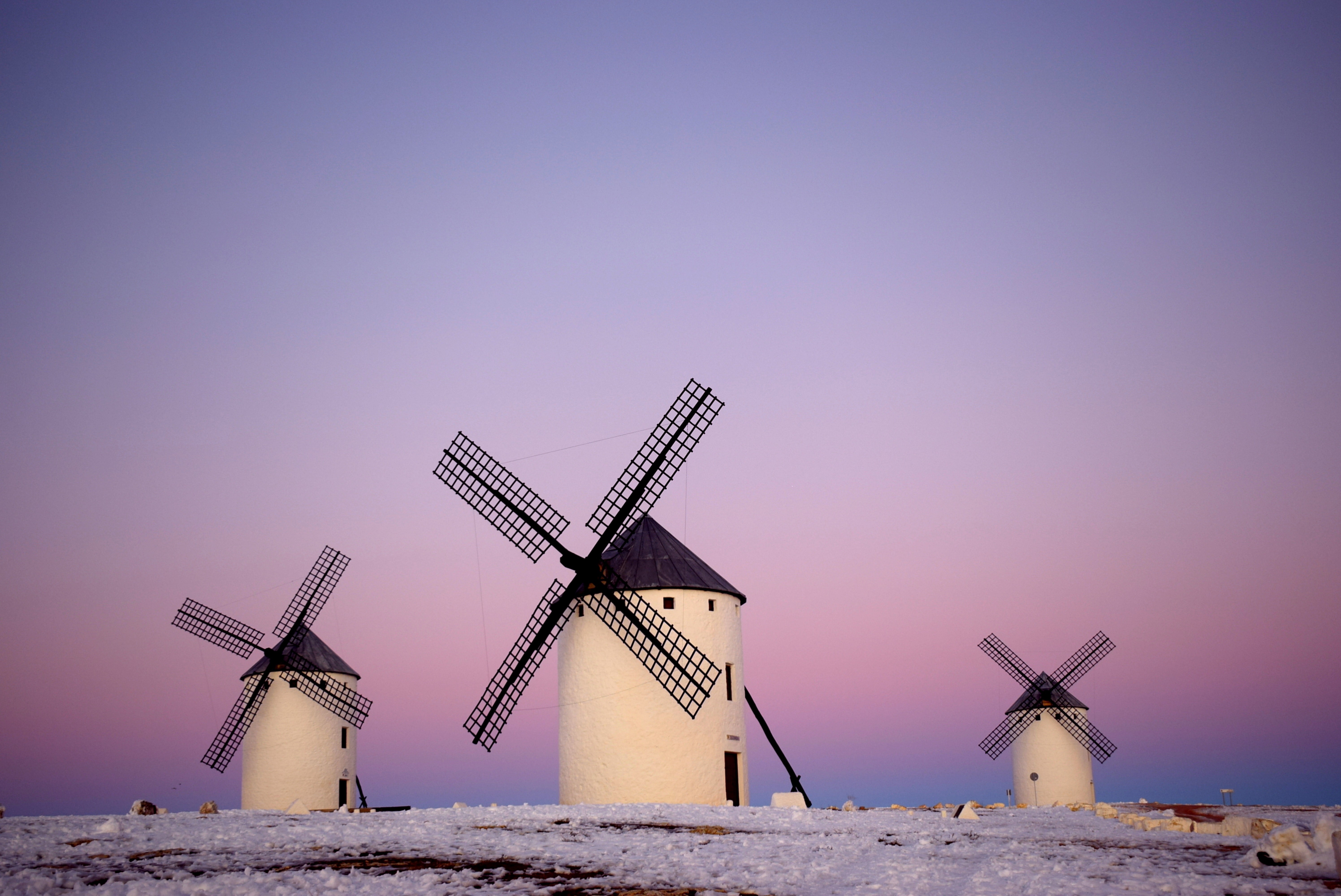 Tres molinos de viento blancos con aspas y tejados negros se alzan en un campo nevado bajo un cielo rosa y púrpura.