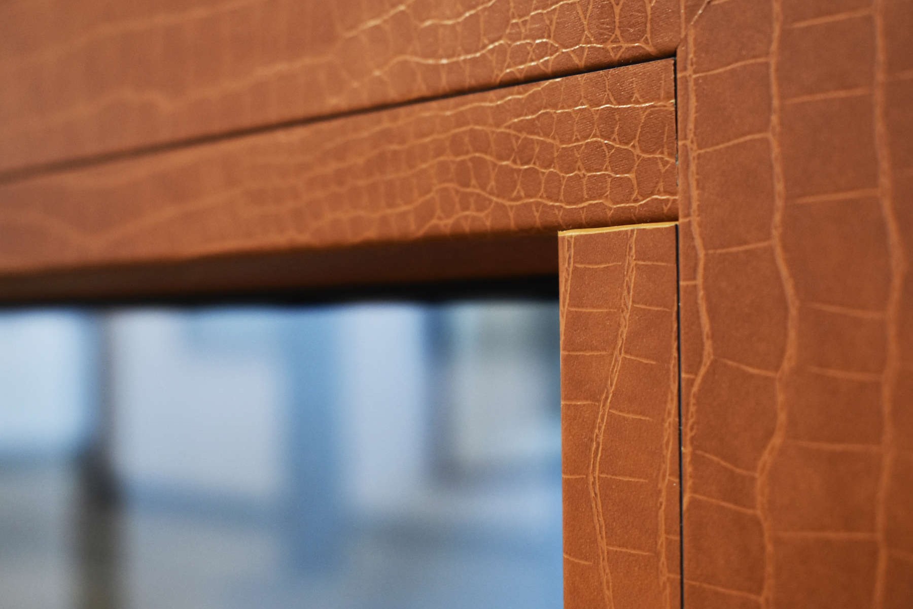 Close-up of a brown wooden frame with a textured surface and a golden border, placed against a blurred background of a glass window.