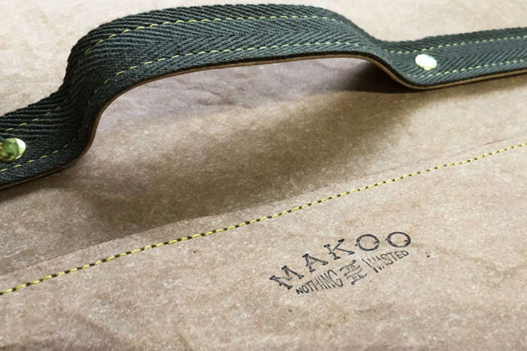 A close-up view of a green leather handle with a yellow stitching on a brown fabric with the words "Mako Nothing Wasted" printed on it.