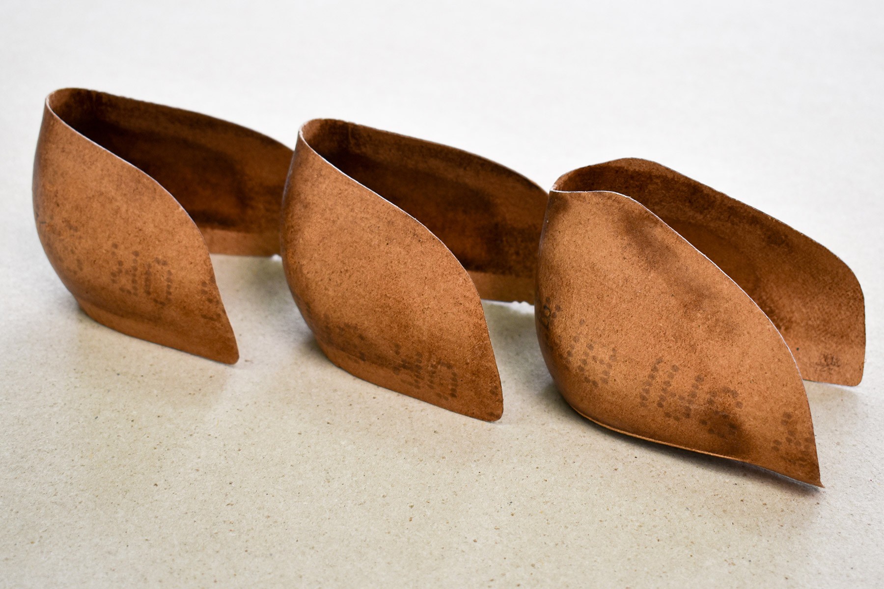 Drei braune, blattförmige Objekte sind auf einer weißen Oberfläche platziert. Die Objekte haben eine strukturierte Oberfläche und scheinen aus einem Material ähnlich wie Papier oder Pappe zu bestehen.
