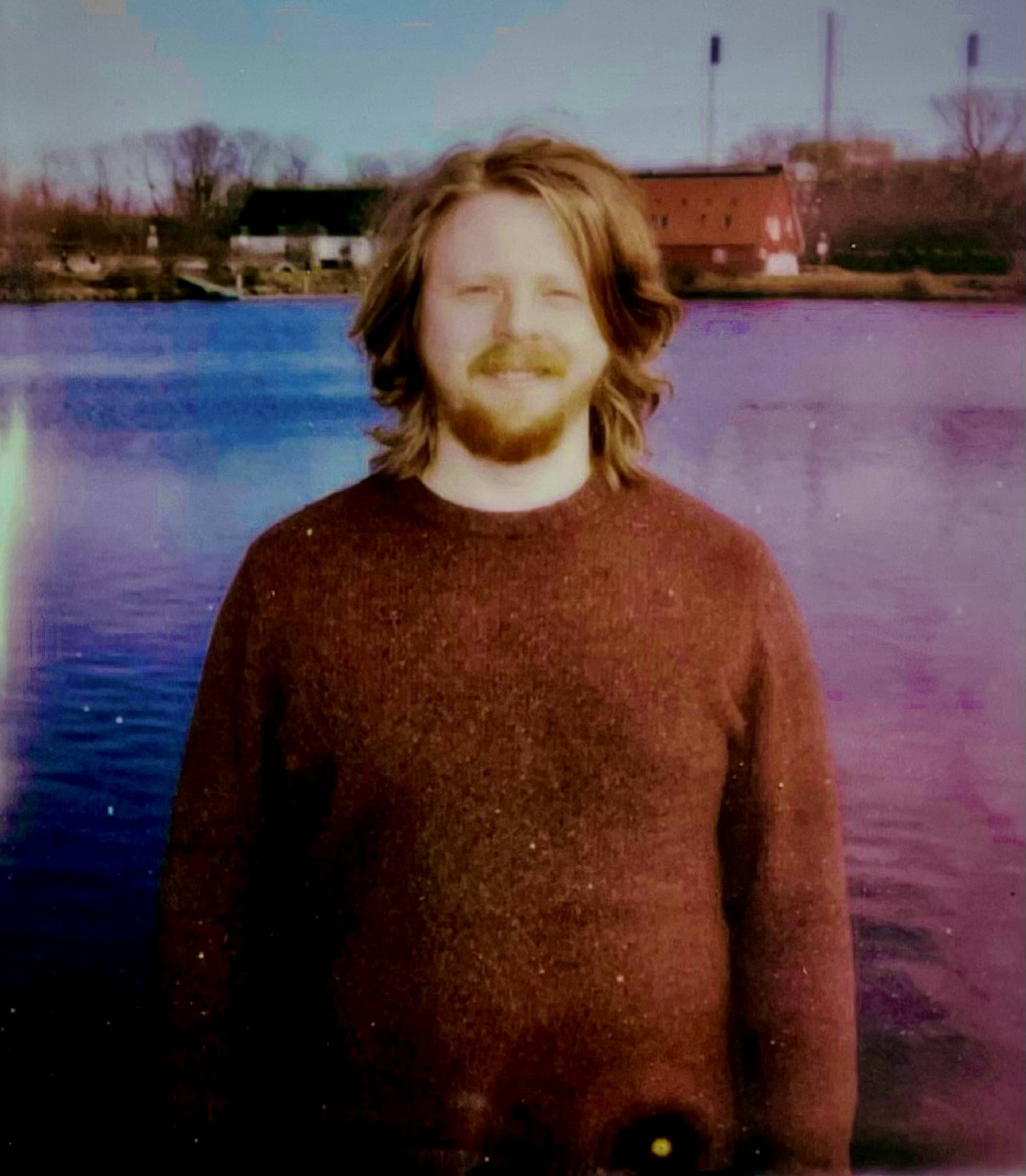 A person with long hair and a beard stands by a calm lake. The background features trees and a red building under a clear sky.