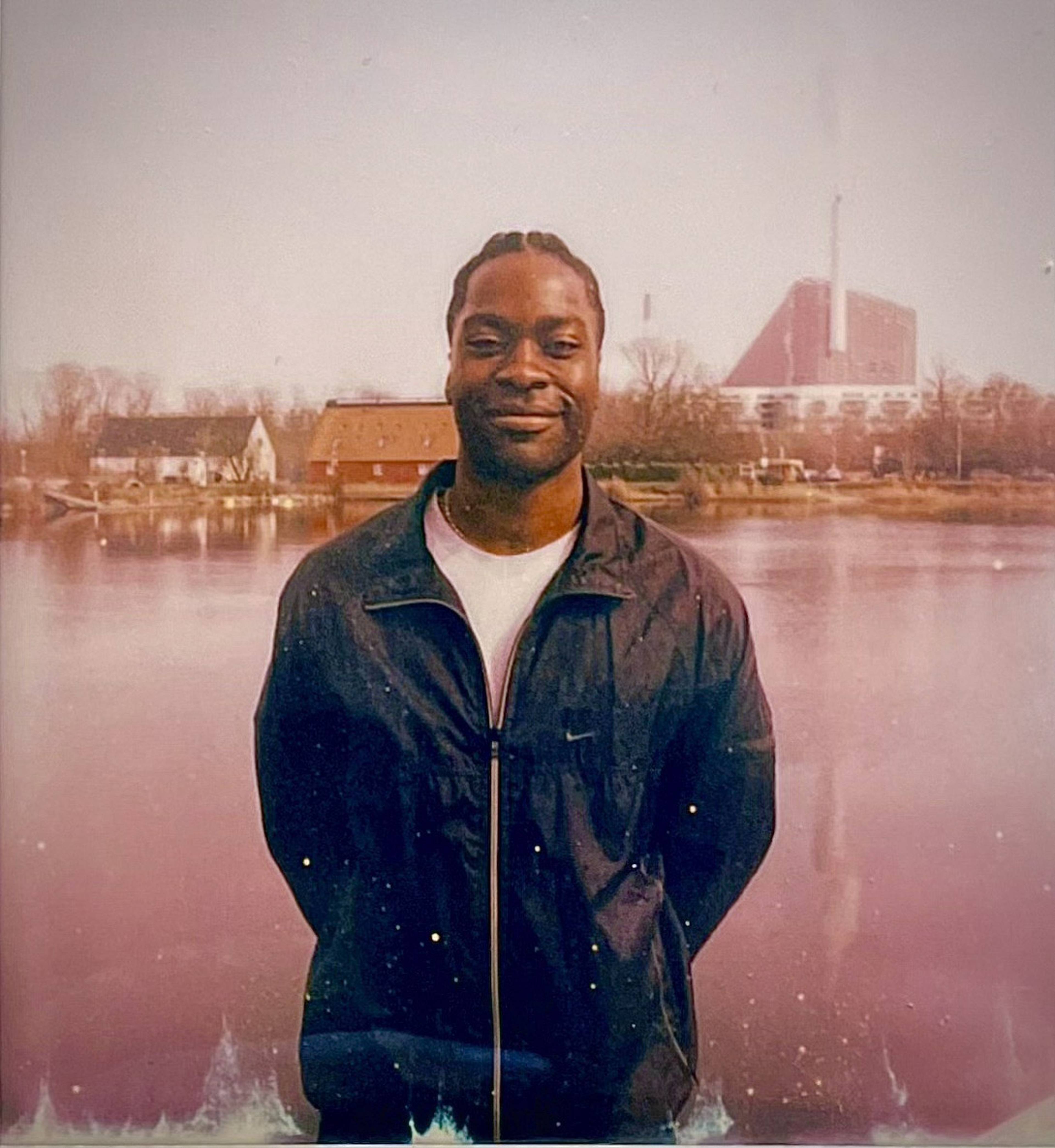 Smiling person in a dark jacket stands by a body of water, with buildings and trees in the foggy background.