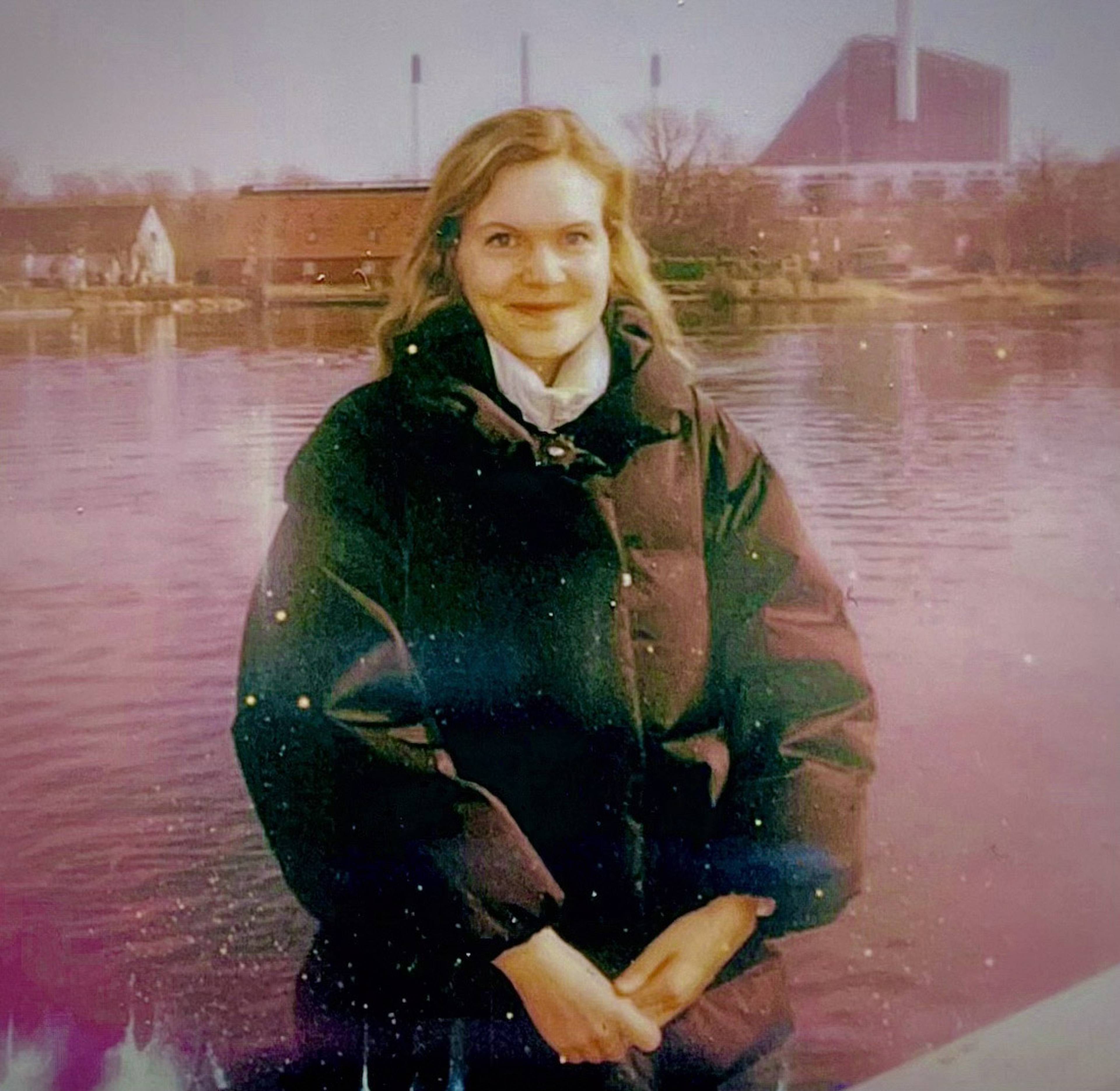 Person in a dark coat standing by a river, with buildings and a factory in the background, under a purplish sky.