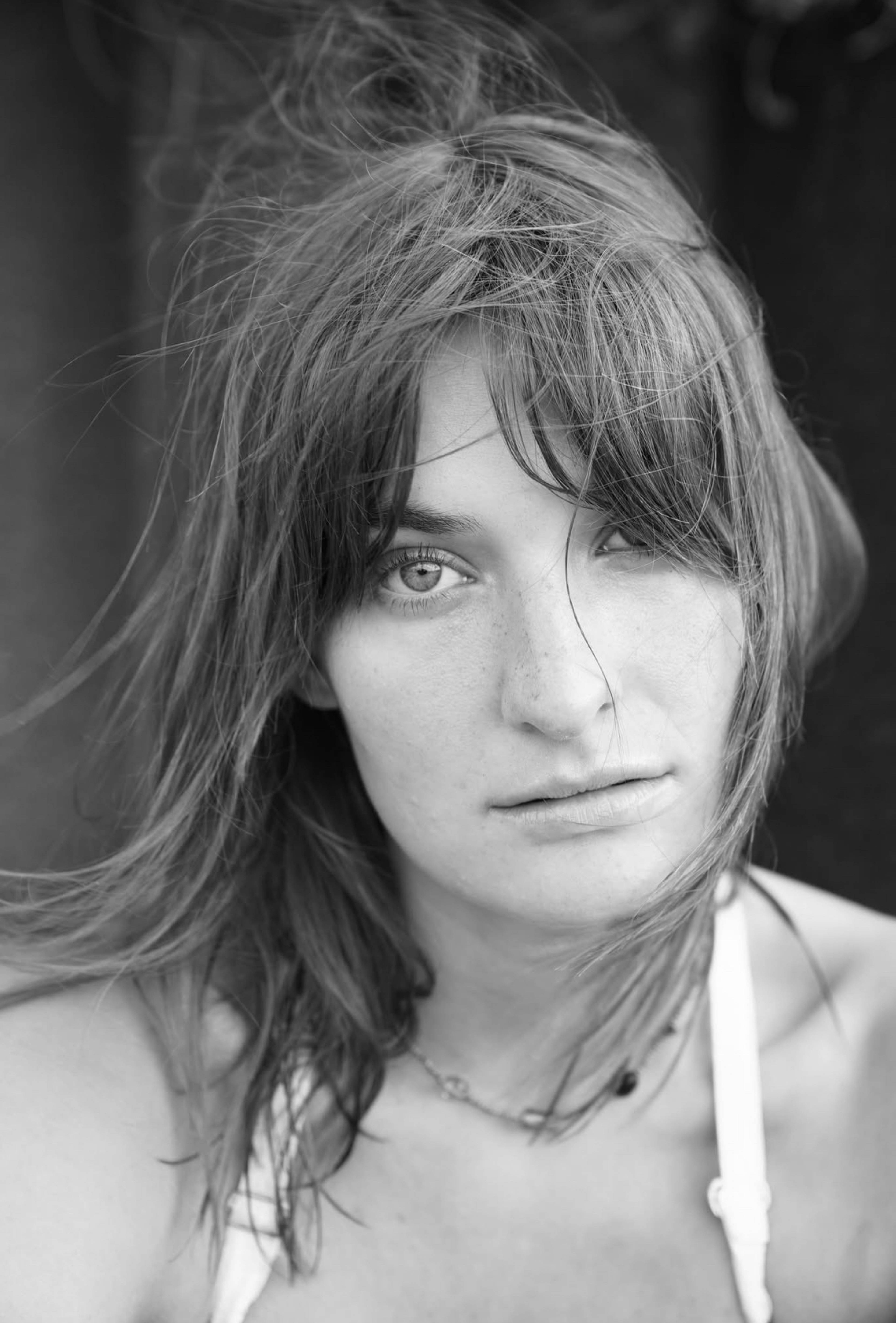 Black and white portrait of a person with tousled hair, wearing a light top, looking intently at the camera.