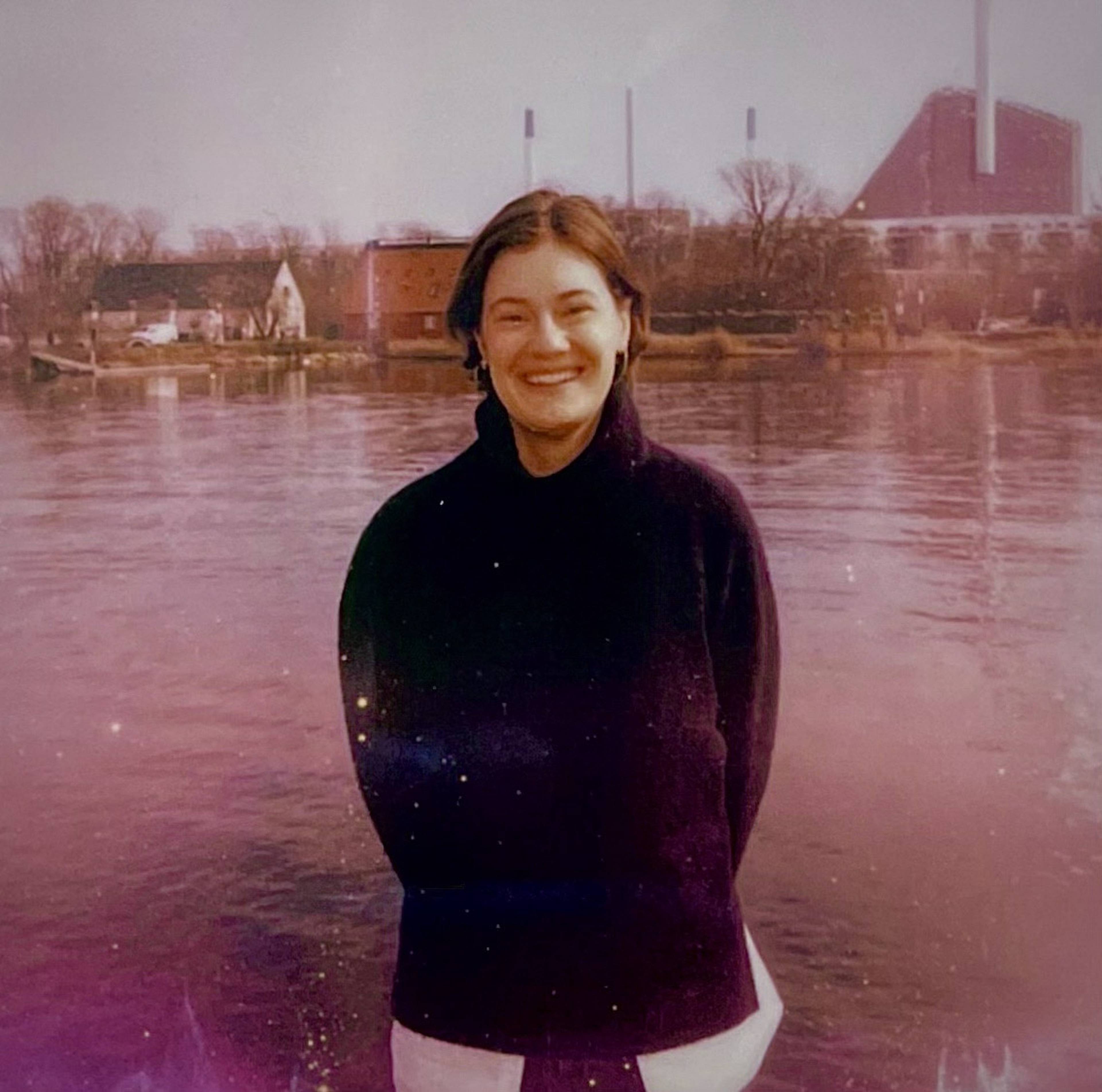 A woman in a dark sweater smiles by a calm river, with industrial buildings and smokestacks in the background on an overcast day.