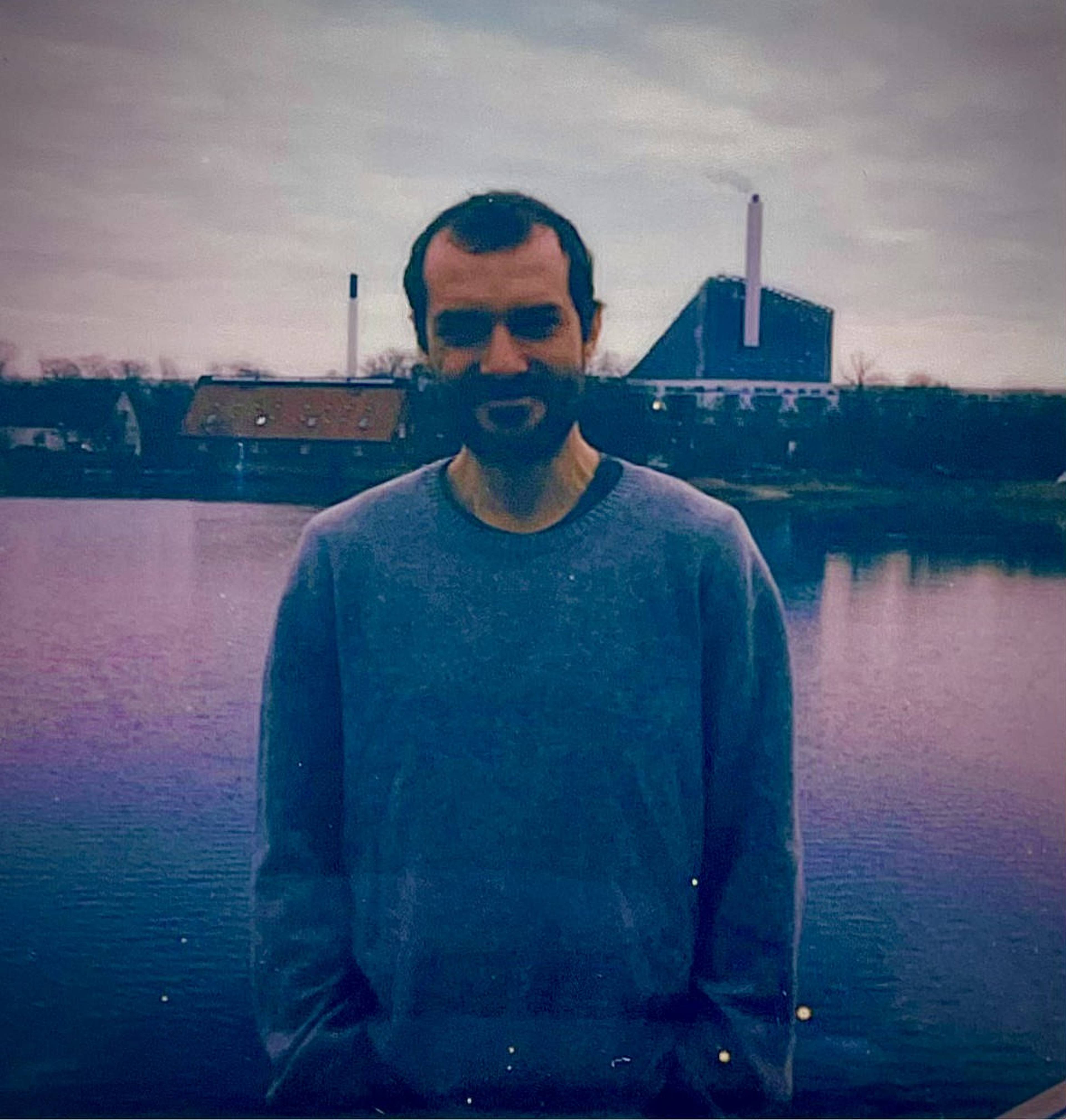 Man in a gray sweater stands by a body of water with an industrial building in the background under a cloudy sky.