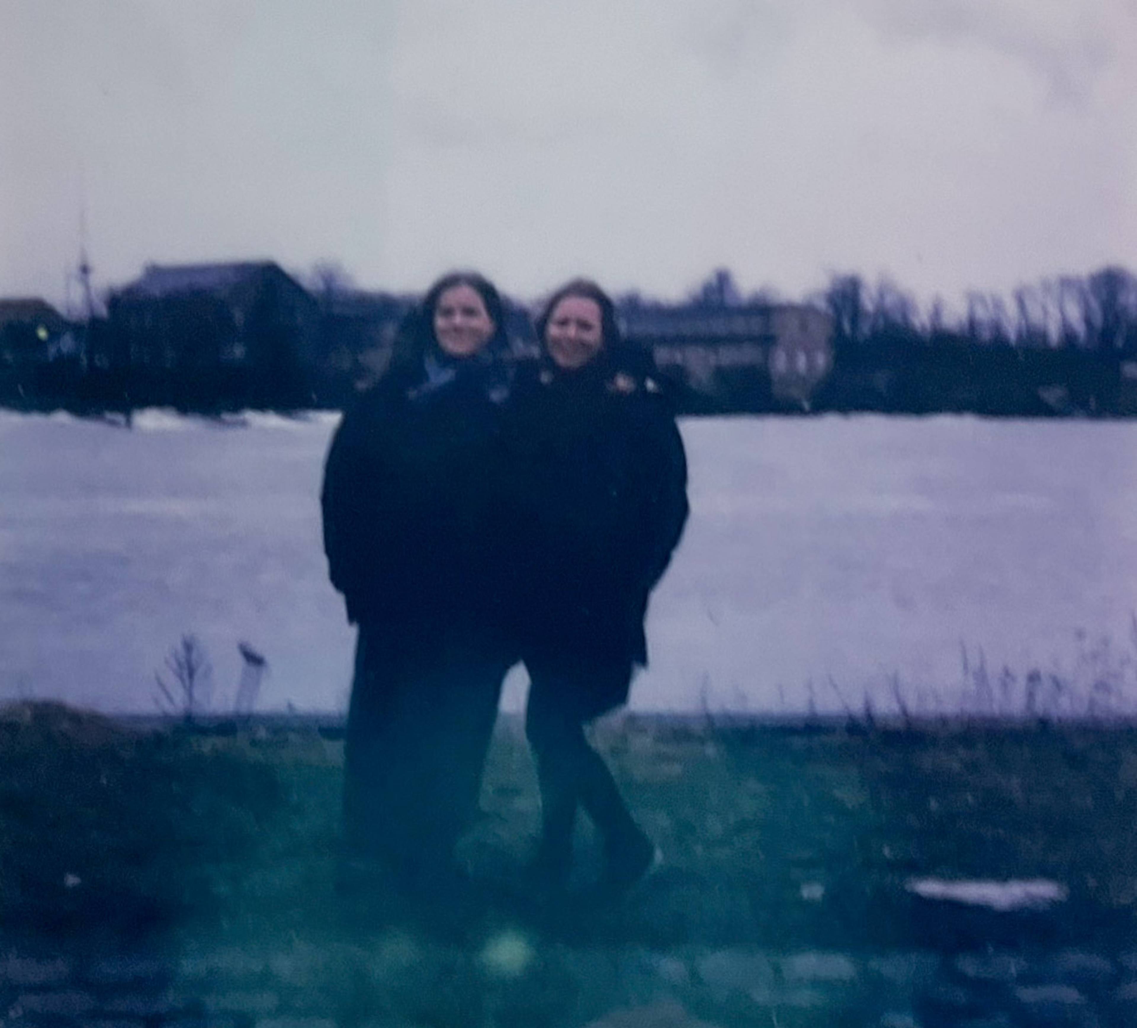 Two people standing together on a grassy area by a river, with trees and buildings in the background on a cloudy day.
