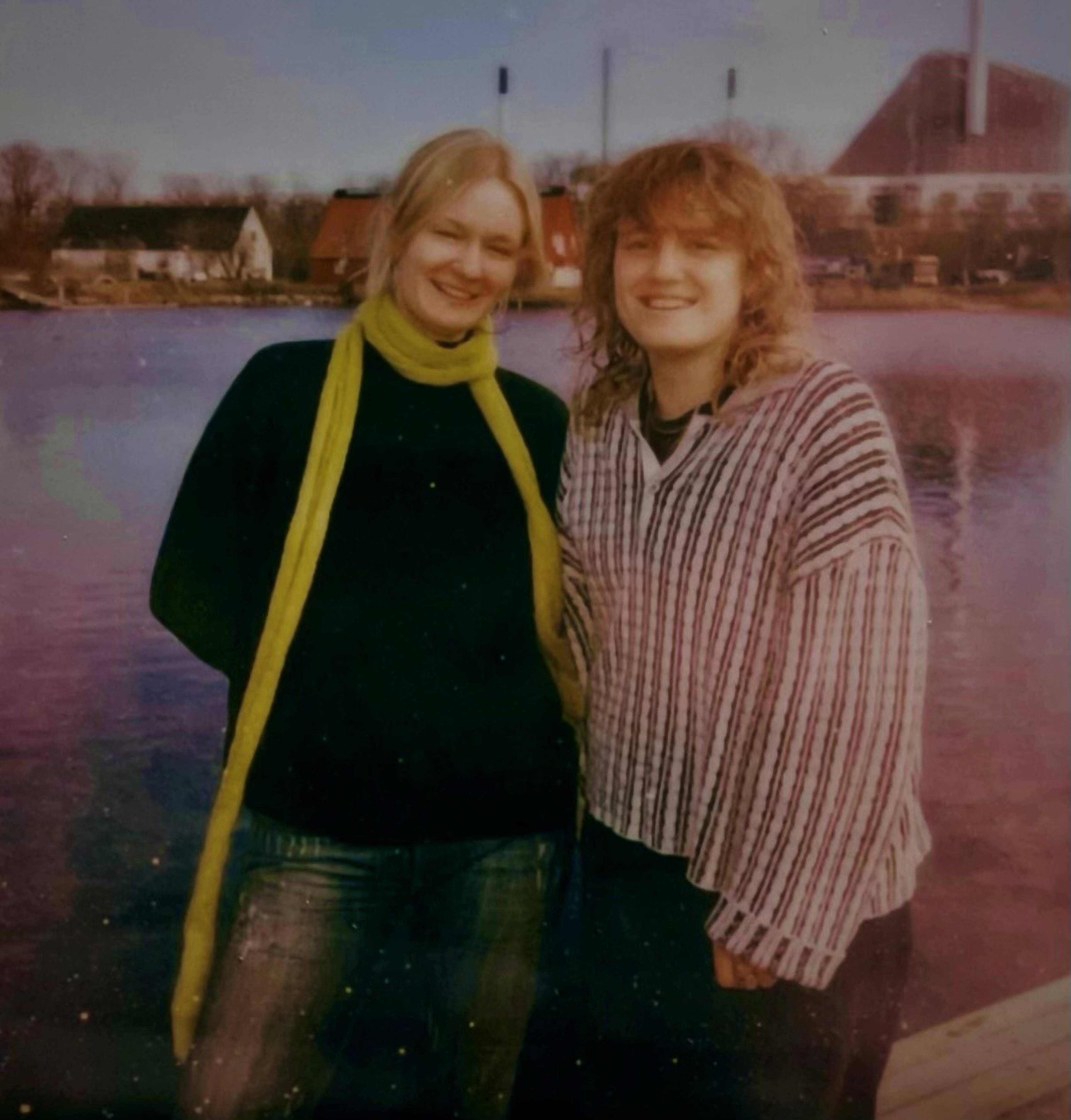 Two people smiling by a waterfront, one wearing a green scarf and the other a striped sweater, with buildings and chimneys in the background.