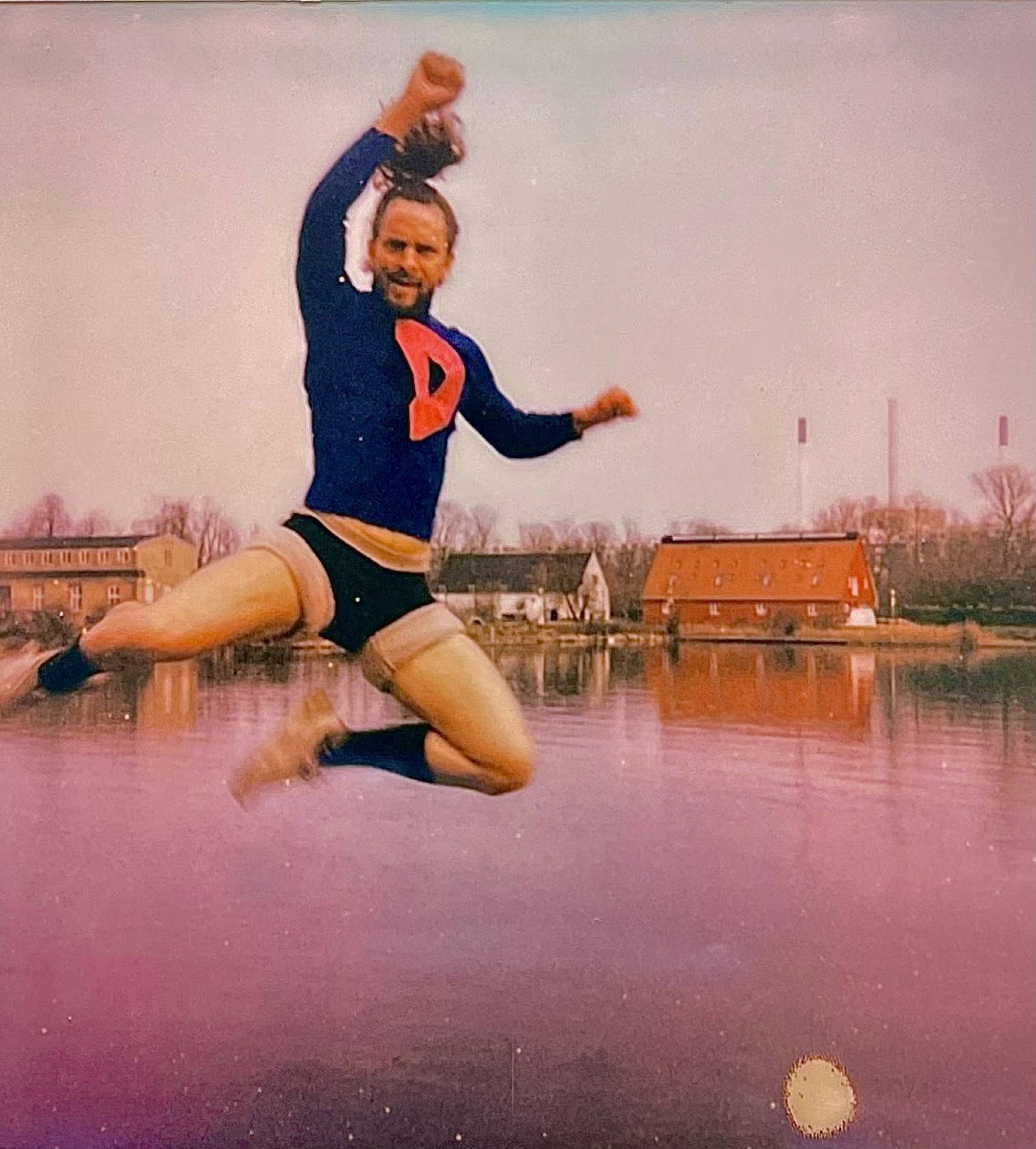 Person in superhero costume joyfully jumps in the air near a lake, with industrial buildings in the background.