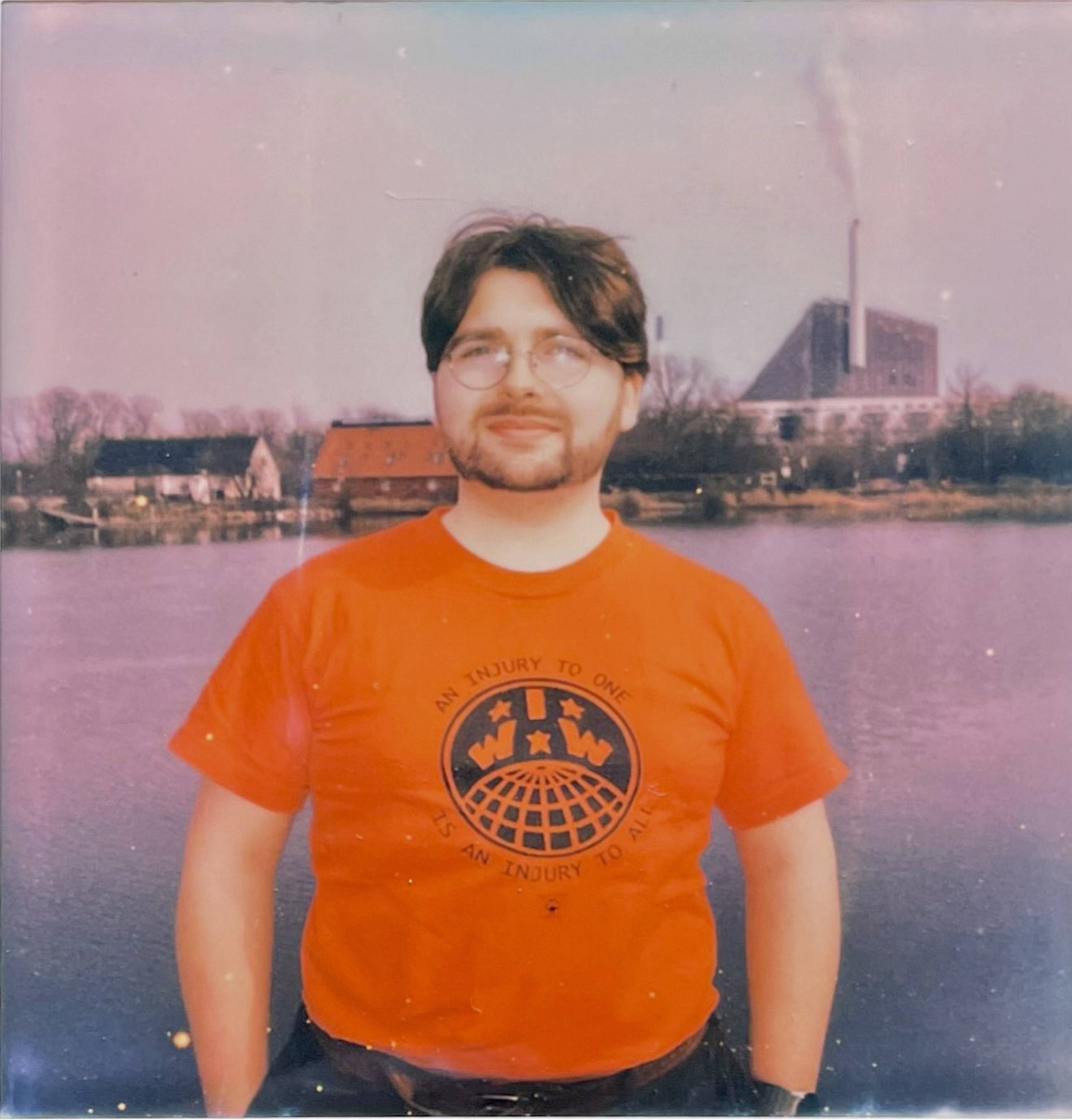 A person wearing a red T-shirt stands by a river with industrial buildings in the background and a hazy sky.