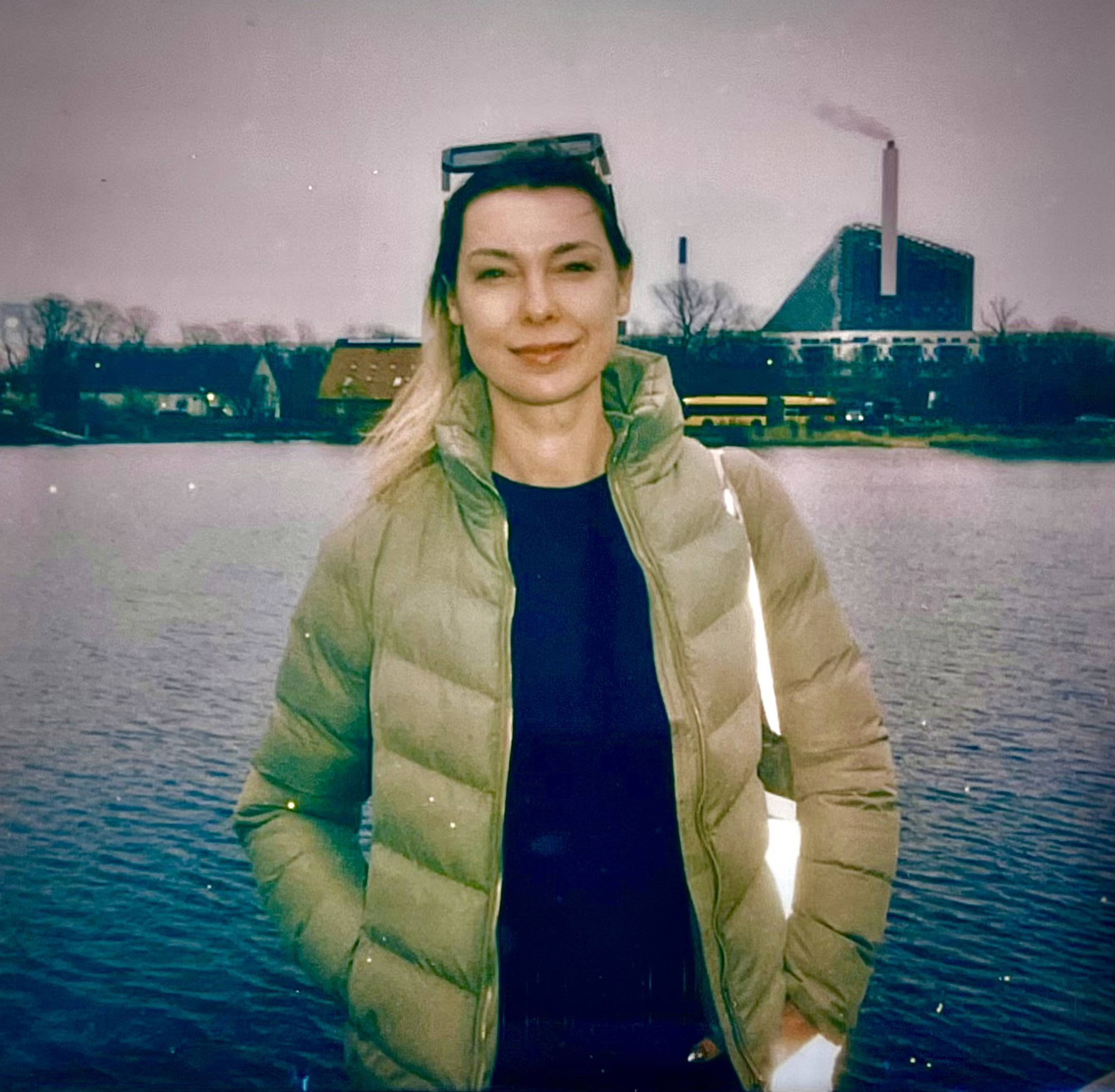 Woman in a green jacket standing by a lake on a cloudy day, industrial building with smoke in the background.