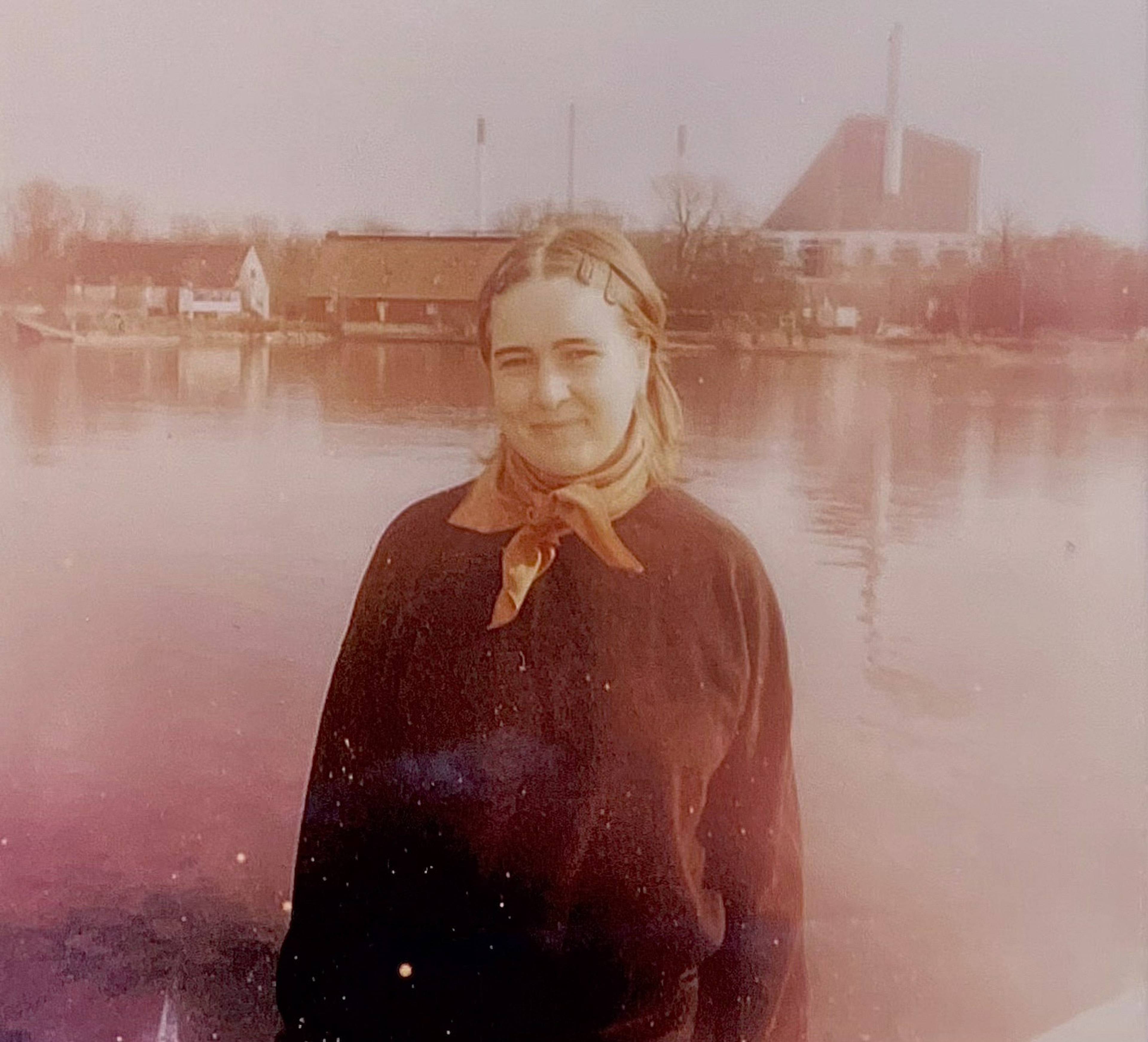 A woman wearing a scarf stands by a calm riverside, with industrial buildings and trees in the background. The image has a vintage tone.