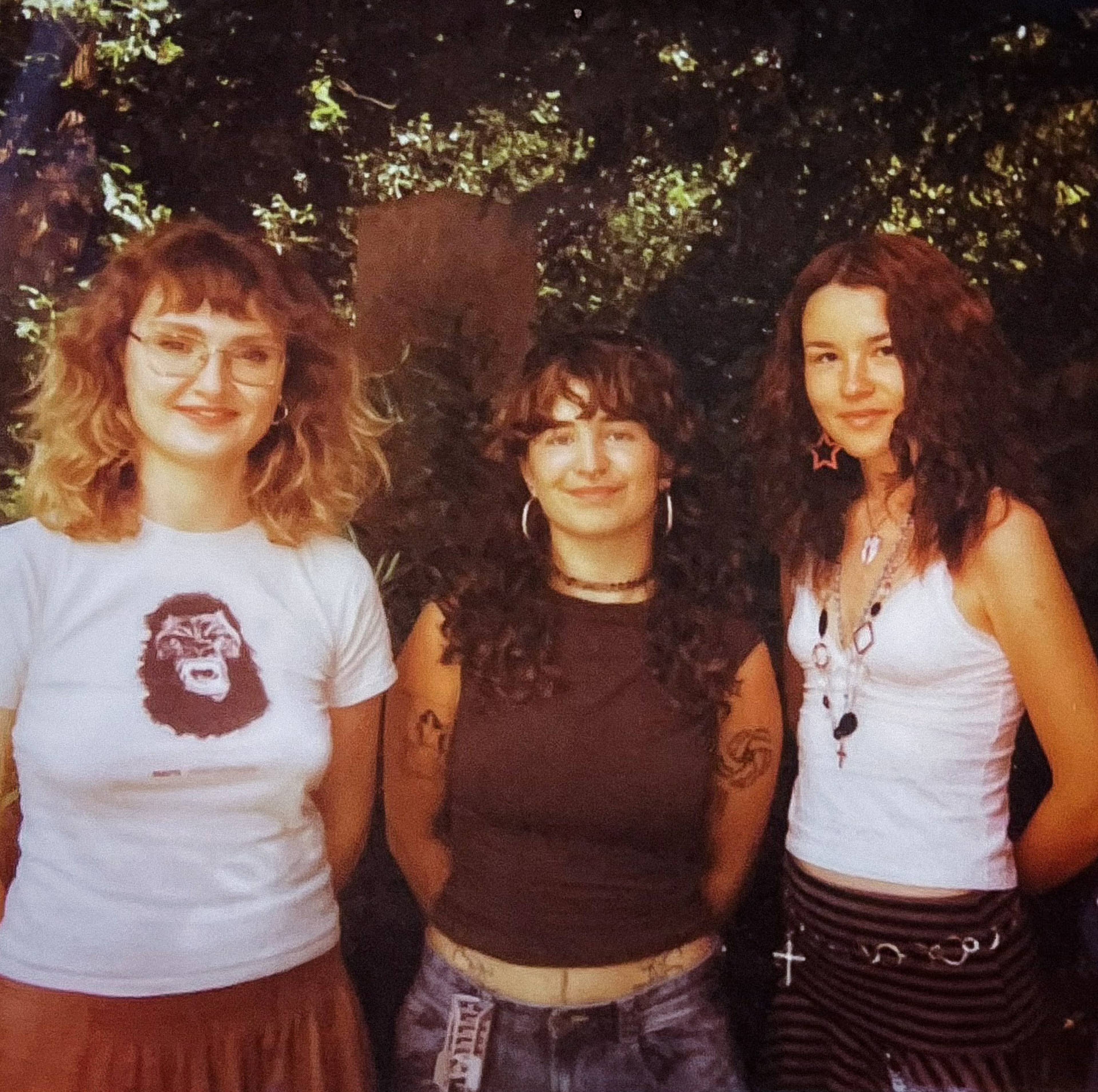 Three young women stand together outdoors, smiling. The first wears a graphic tee, the second a dark tank top, and the third a white tank with accessories.