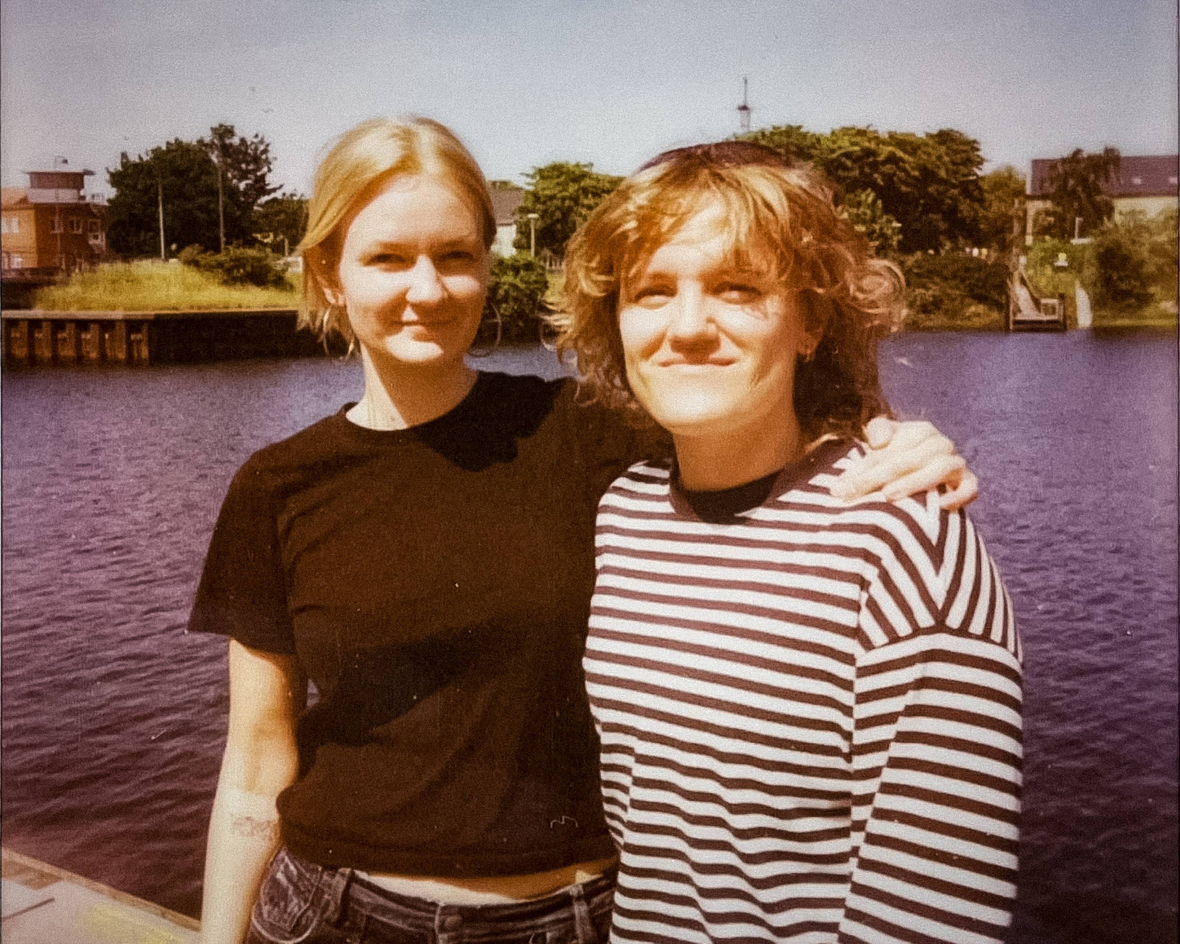 Two people smiling by a riverside, with one wearing a black shirt and the other in a striped shirt. Trees and buildings are visible in the background.