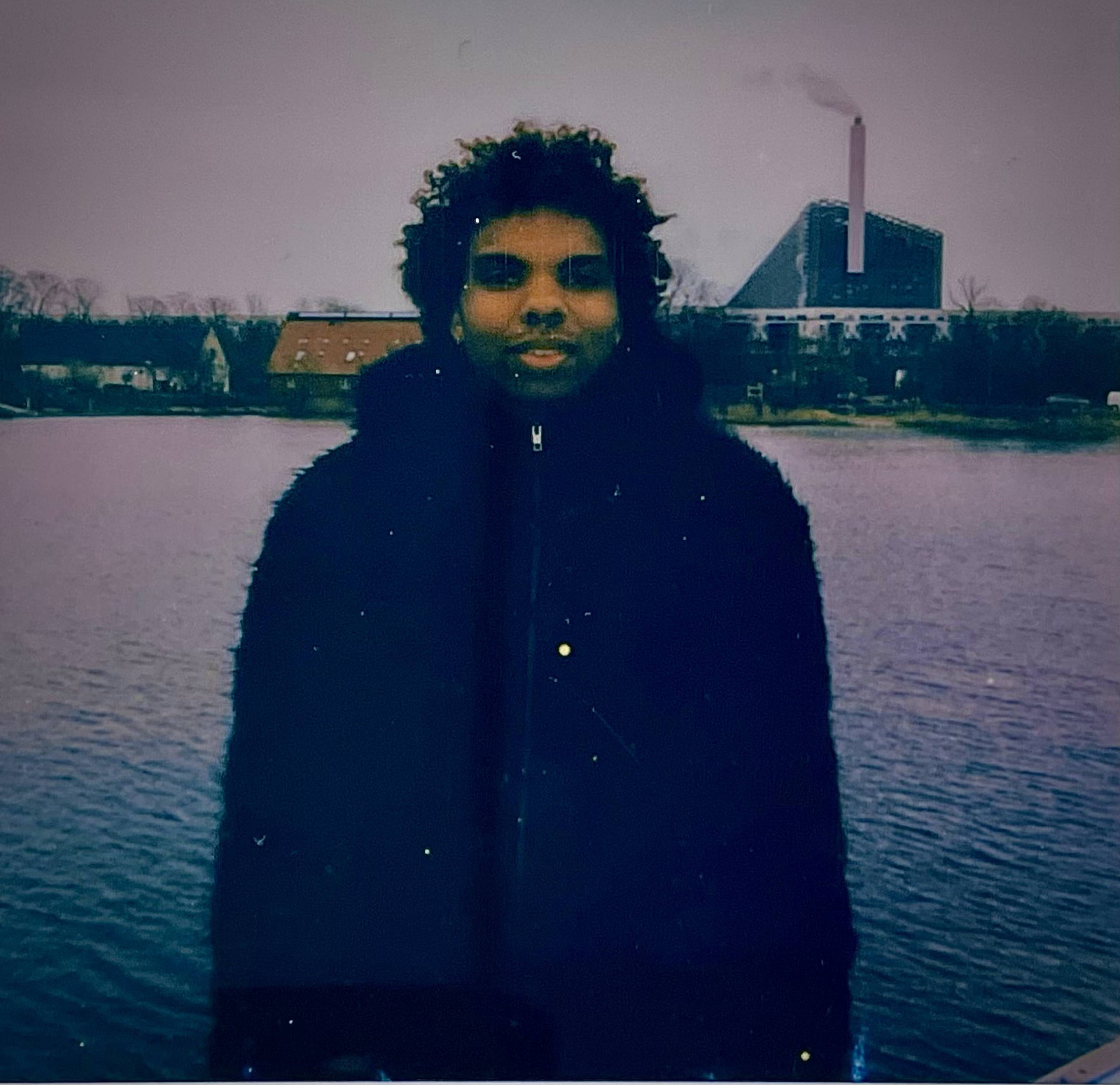 Person standing by a body of water, with a large building emitting smoke in the background on a cloudy day.