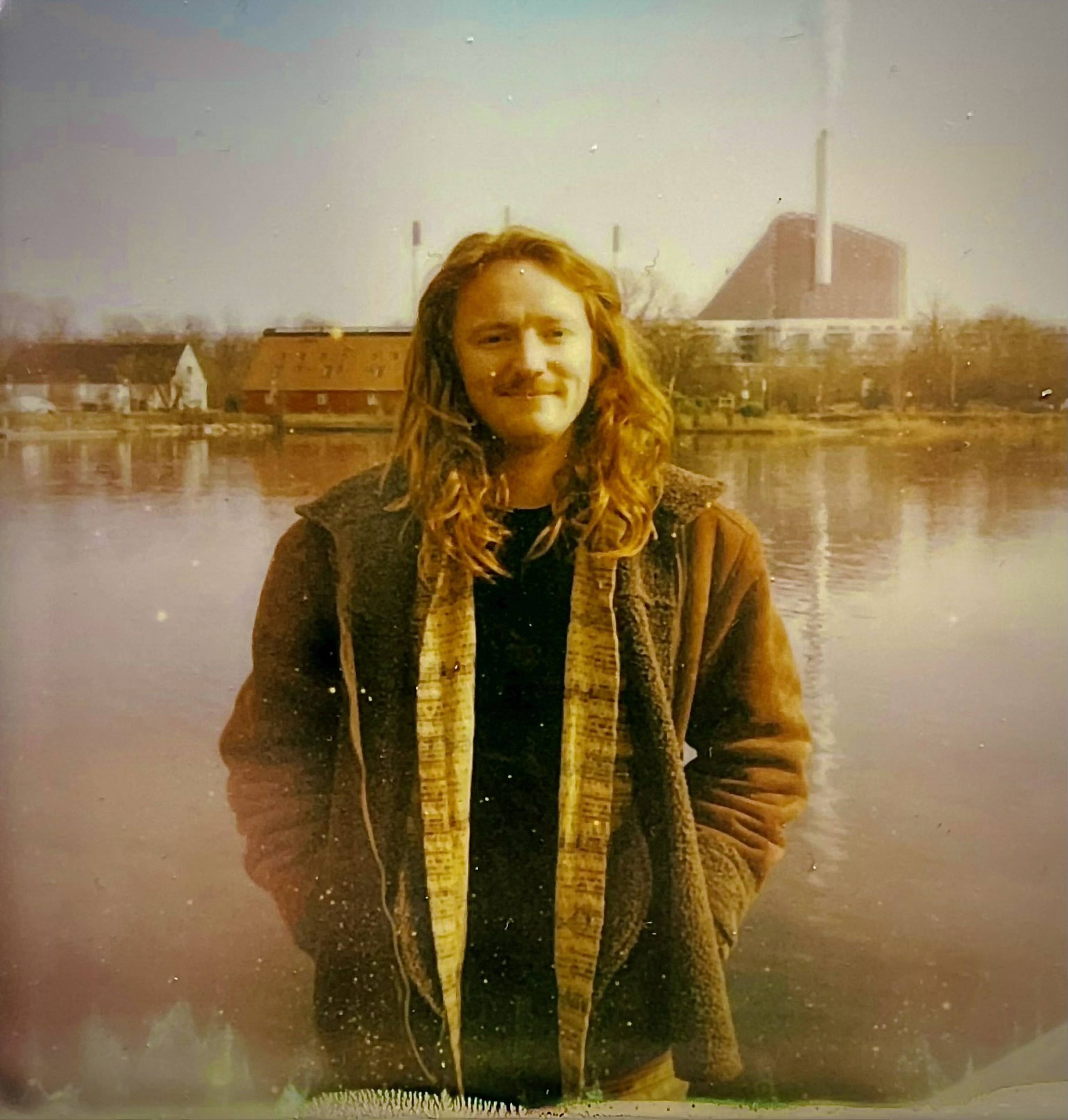 Person with long hair stands by a calm lake, wearing a brown jacket. A large industrial building is visible in the background.