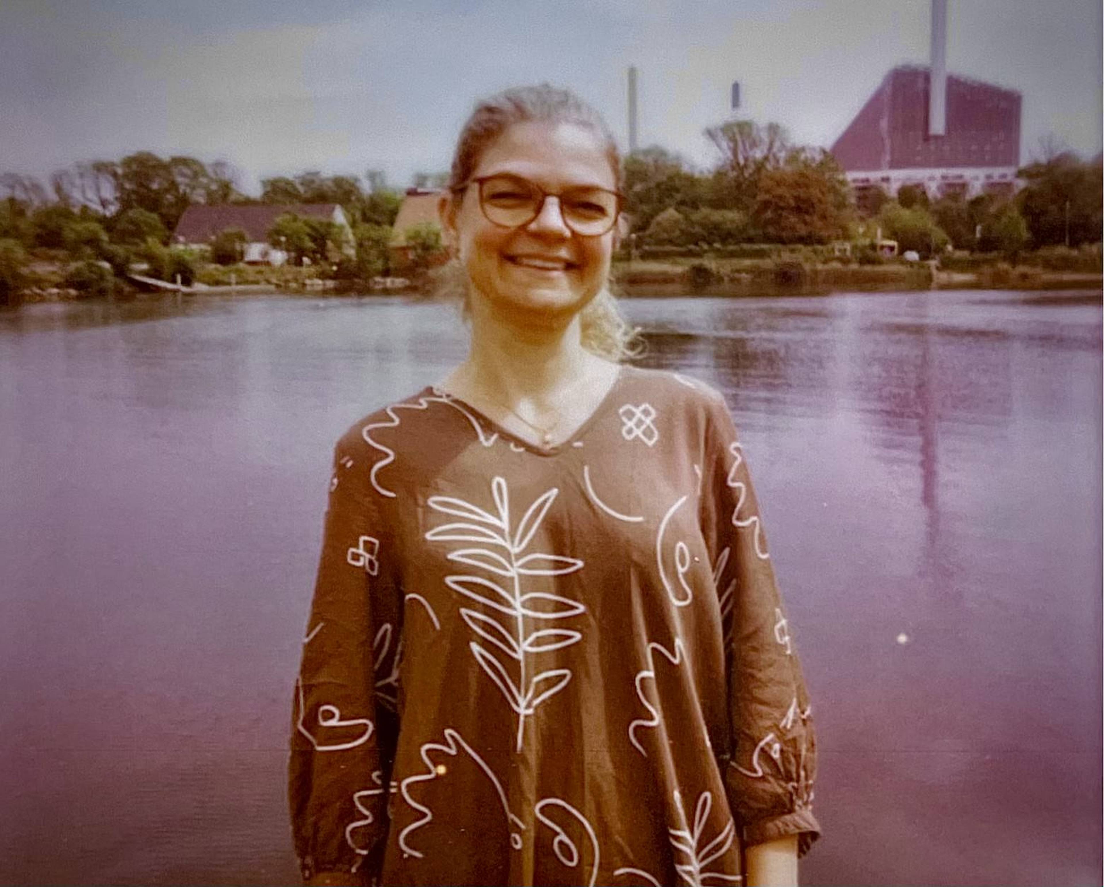 Woman smiling by a lake, wearing glasses and a patterned dress, with trees and industrial buildings in the background.