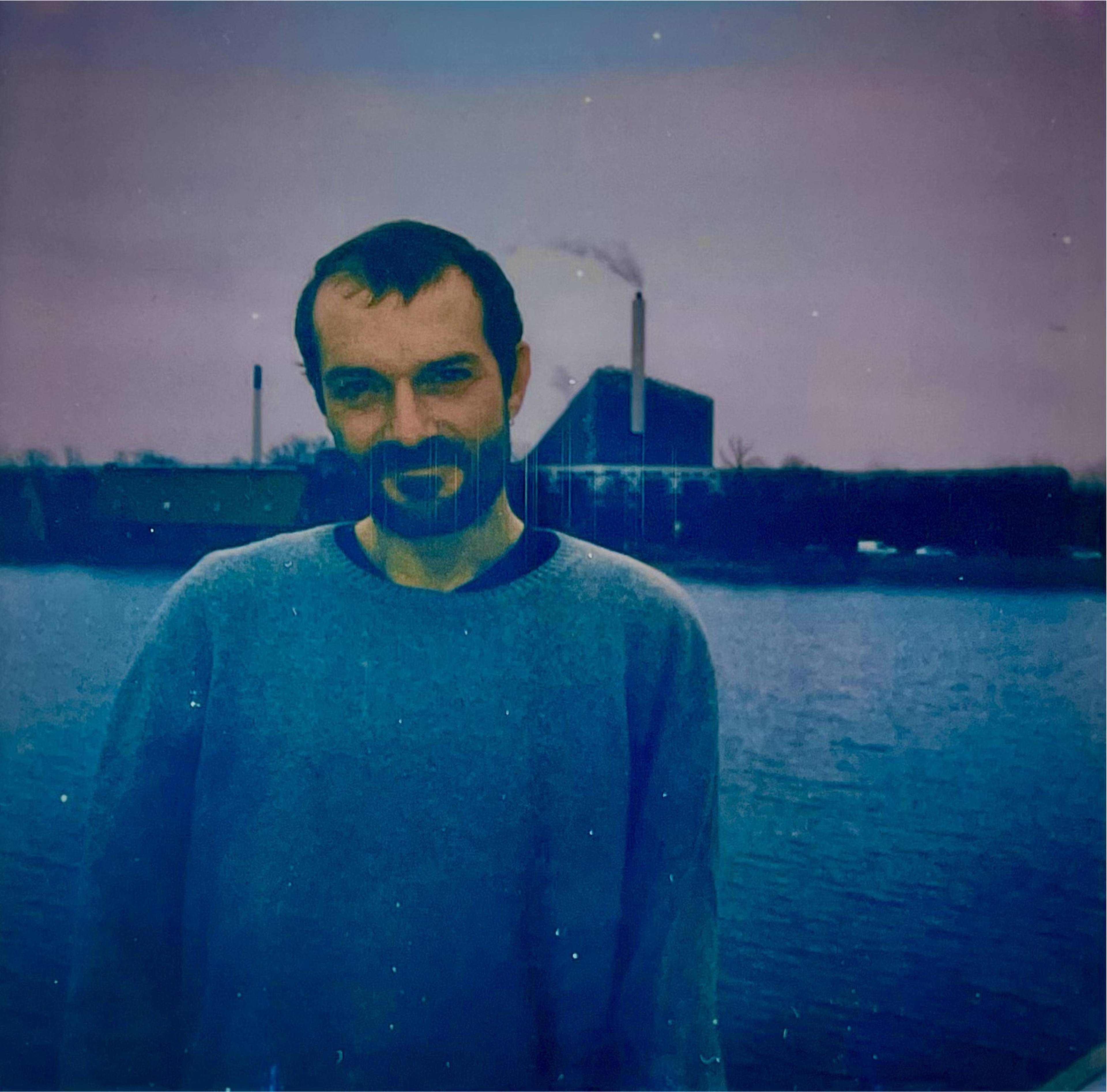 A man with a beard and a sweater stands near water with an industrial building emitting smoke in the background, under a dusky sky.