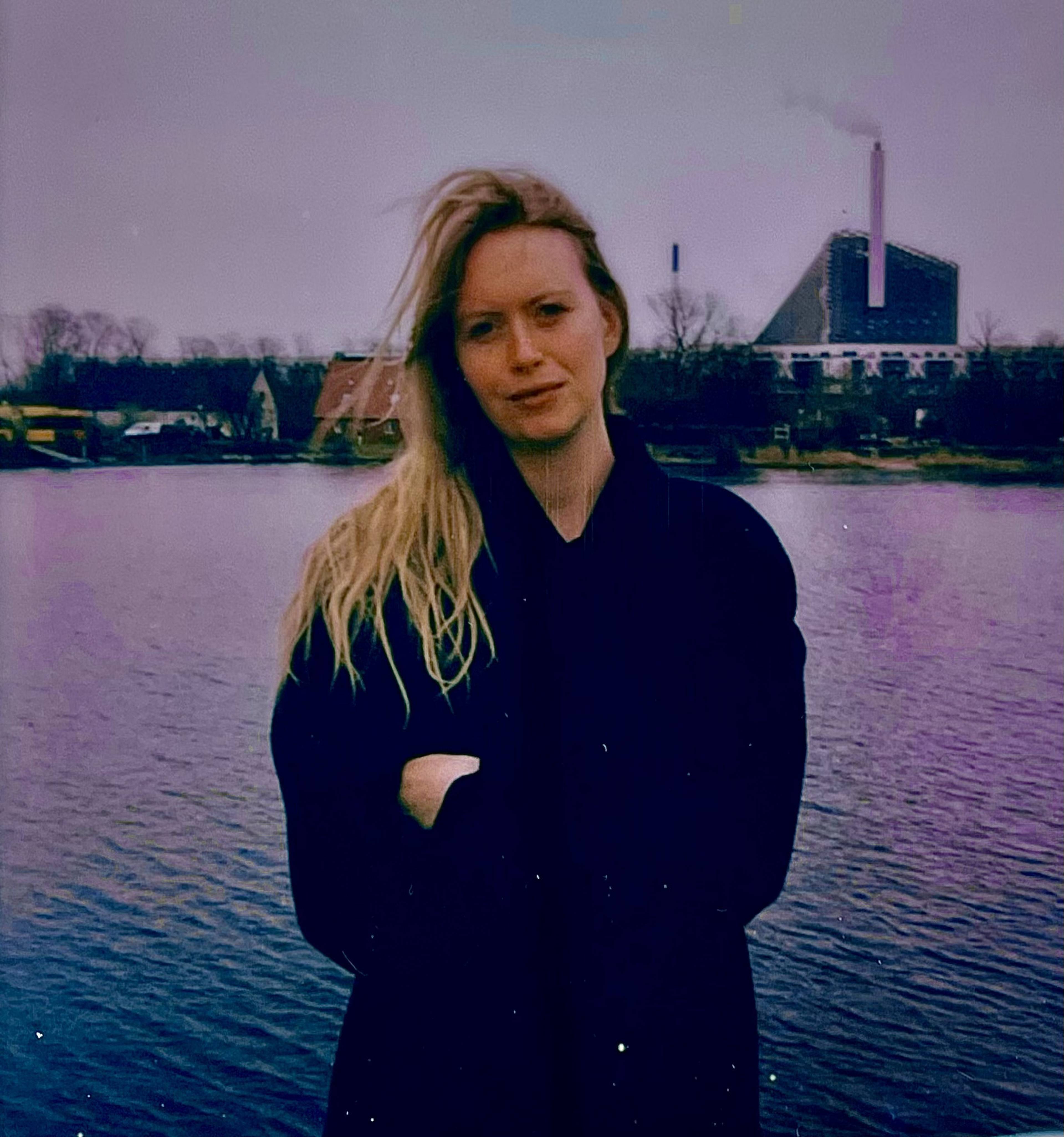 Woman with long blonde hair stands by a lake, arms crossed, with an industrial building in the background on a cloudy day.