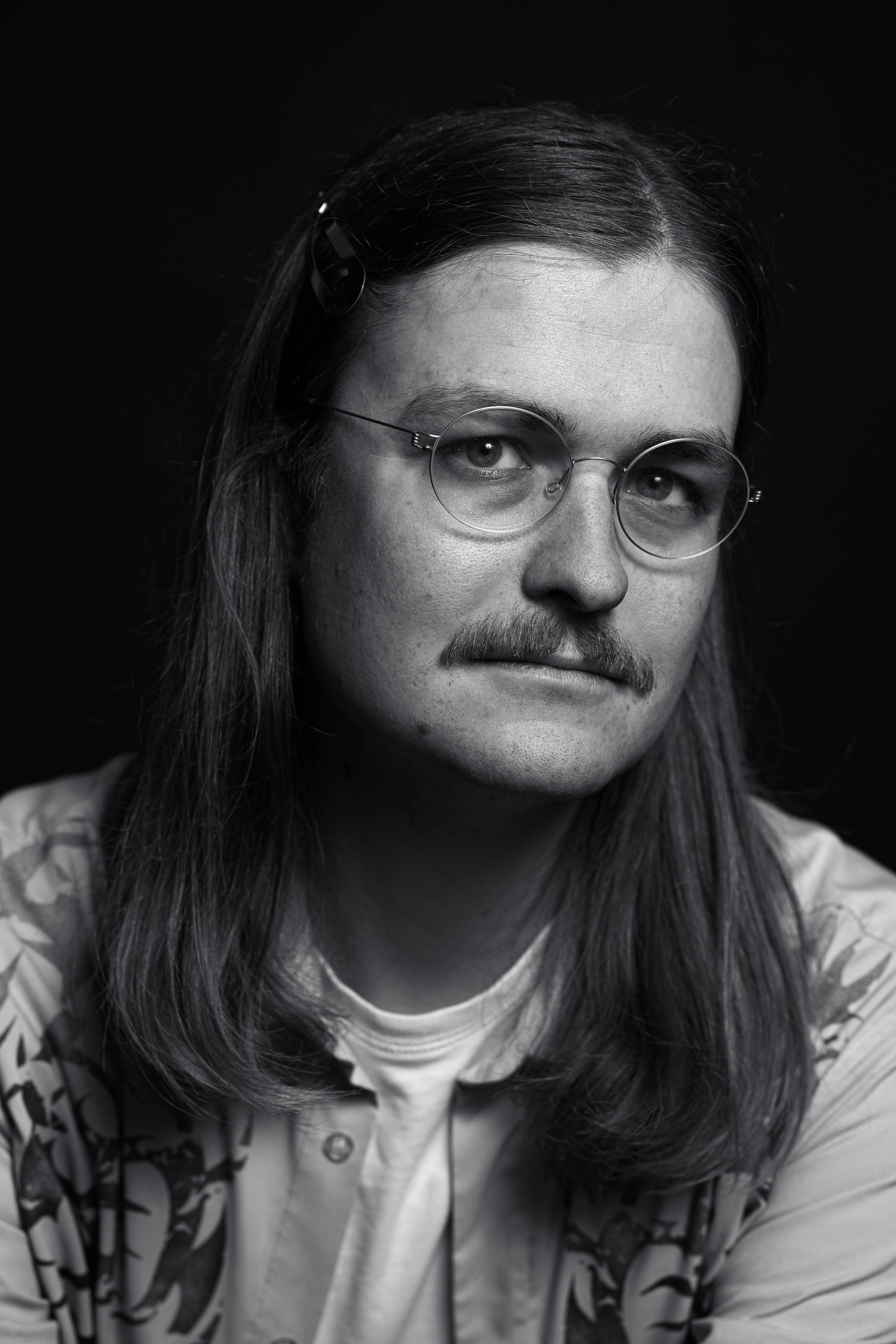 Black and white portrait of a person with long hair, glasses, and a mustache, wearing a patterned shirt against a dark background.
