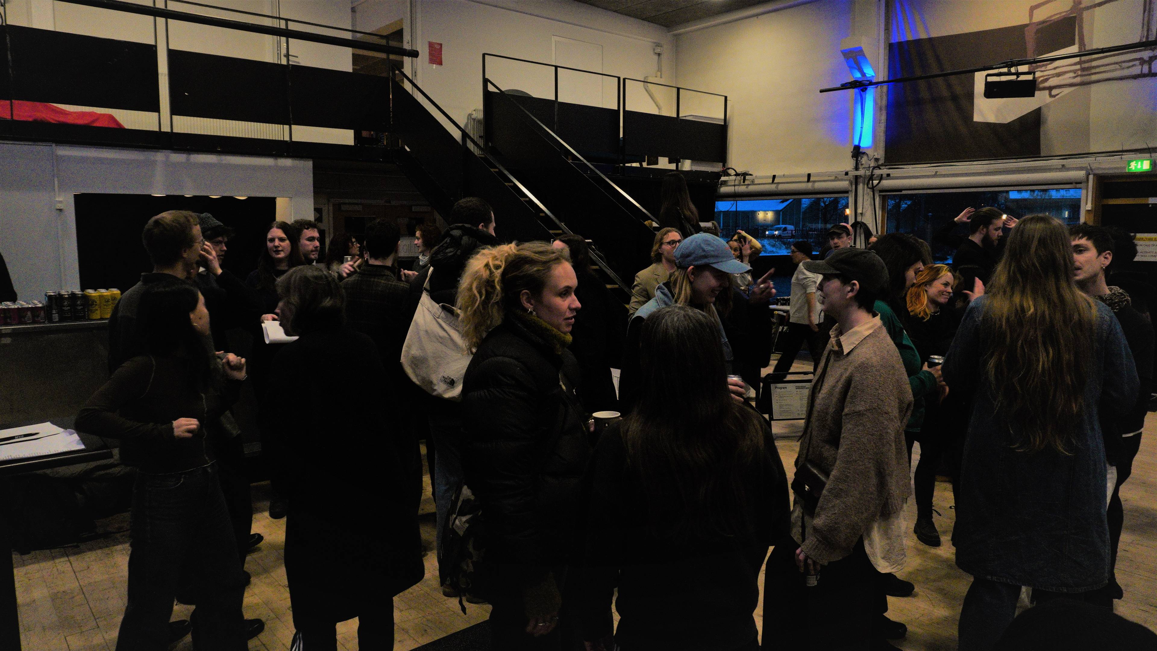 A group of people gathering indoors, engaging in conversation and socializing. Stairs and seating area are visible in the background.