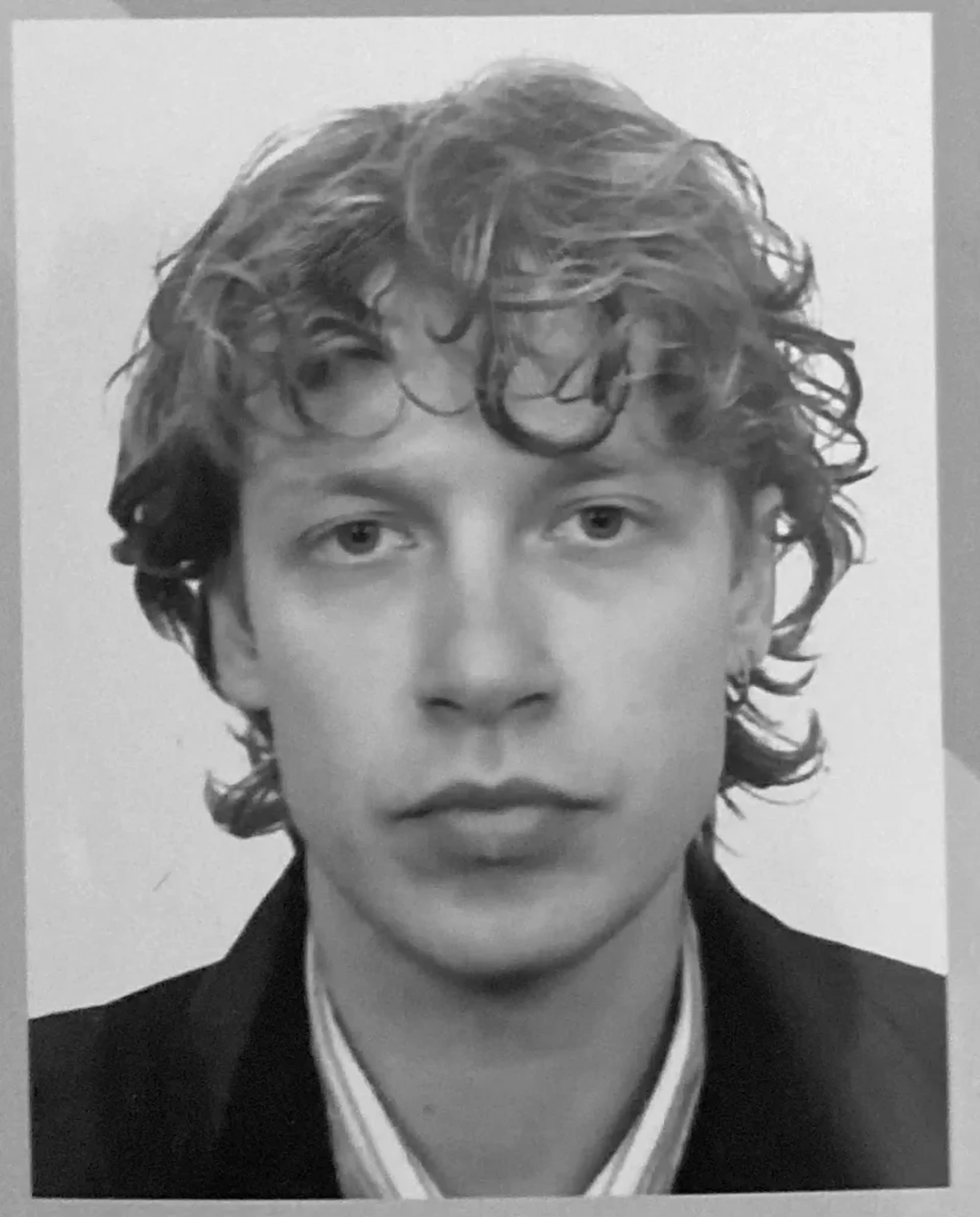 Black and white portrait of a person with curly hair, wearing a jacket and striped shirt, looking directly at the camera.