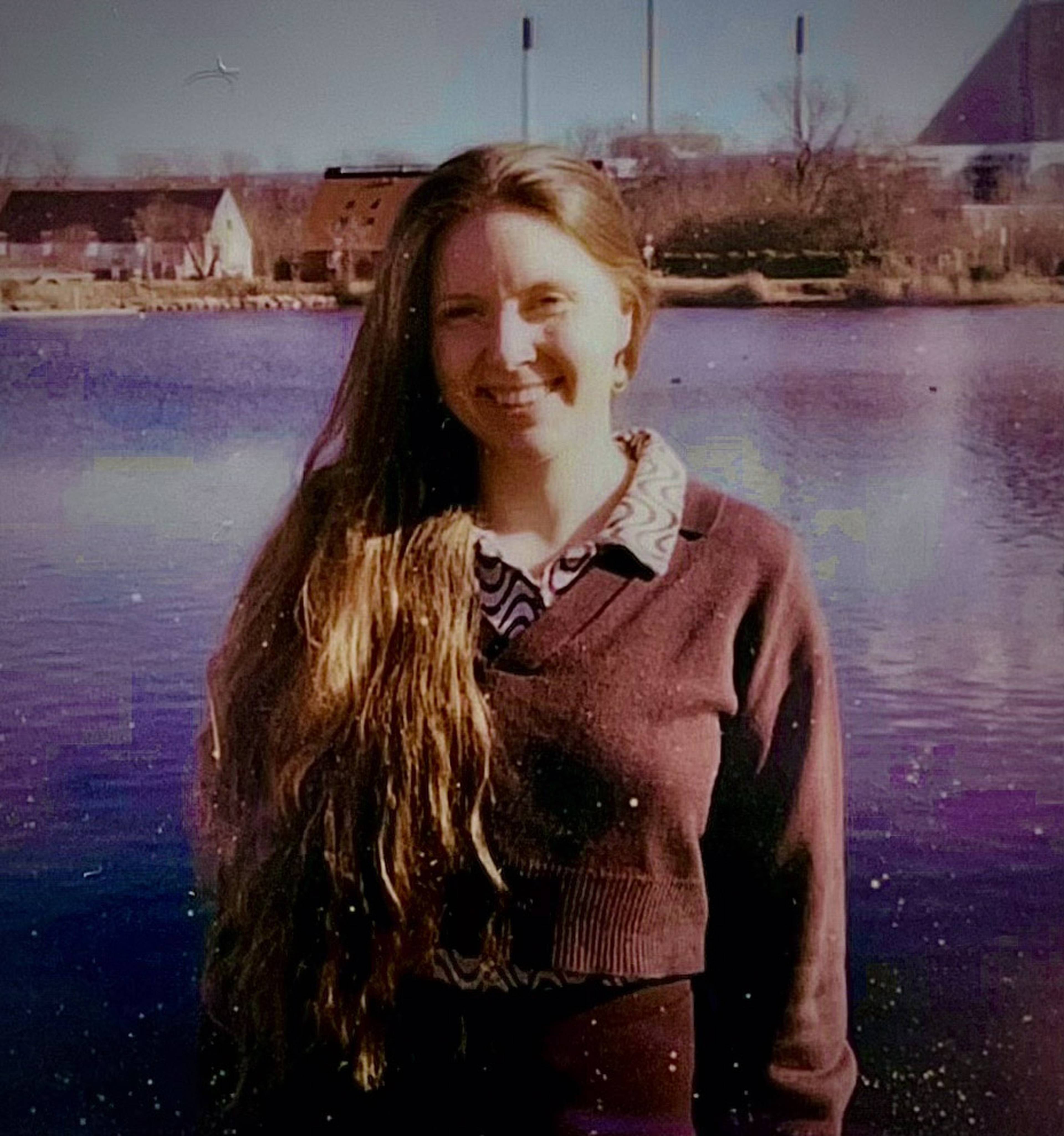 Smiling woman with long hair stands by a reflective lake, industrial buildings and bare trees in the background.