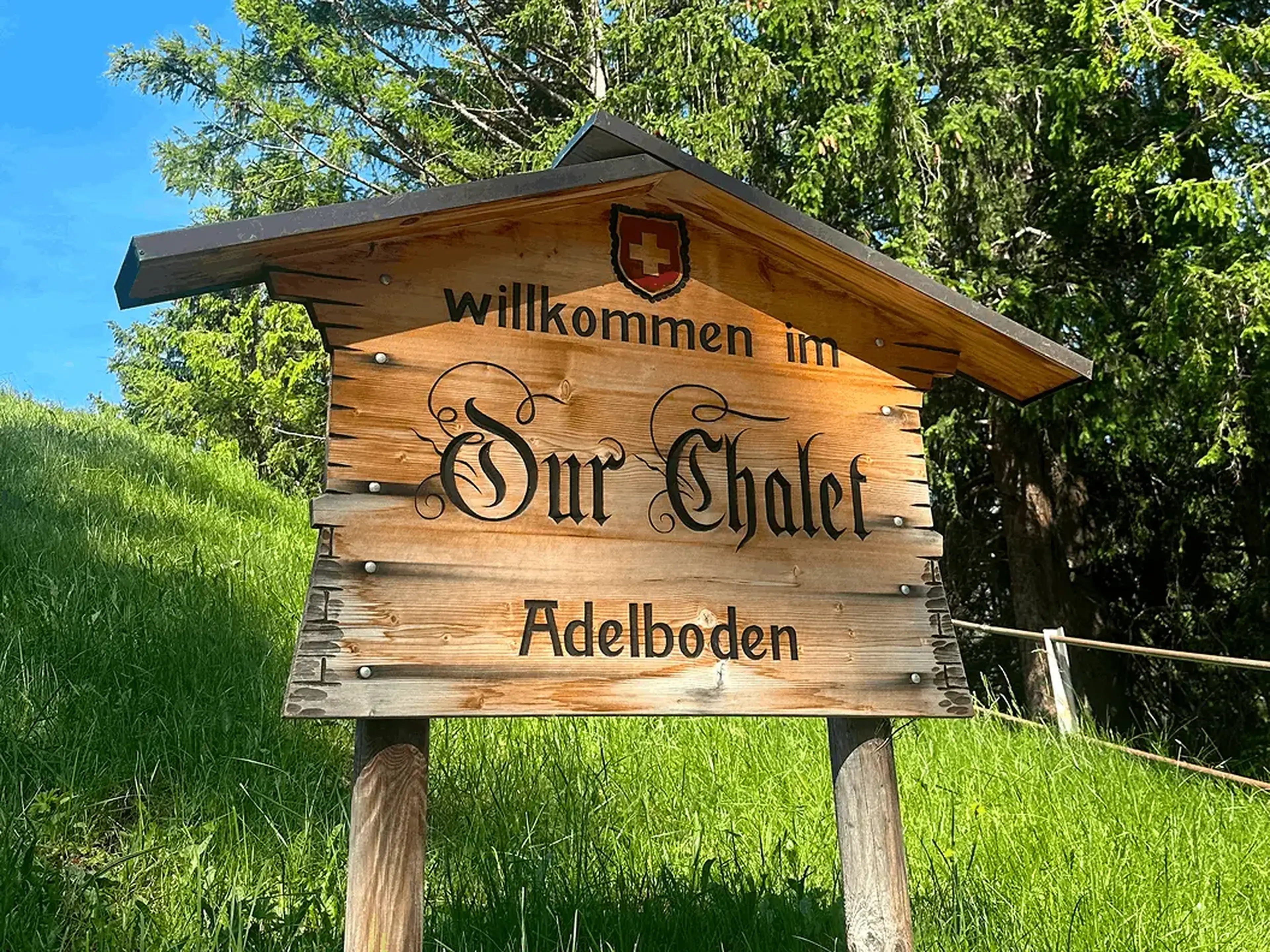 A wooden sign with "Willkommen im Our Chalet Adelboden" stands on green grass, backed by lush trees.
