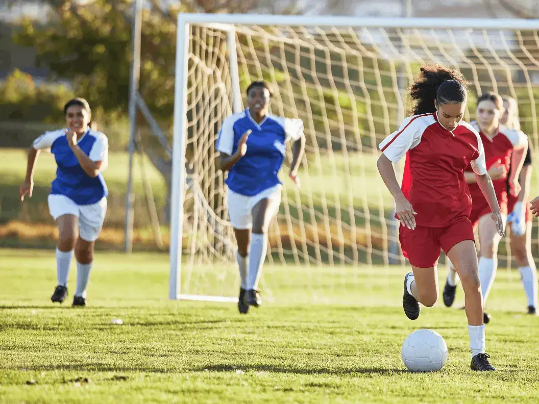A soccer player in red dribbles a ball toward the goal, pursued by players in blue jerseys on a sunny field.