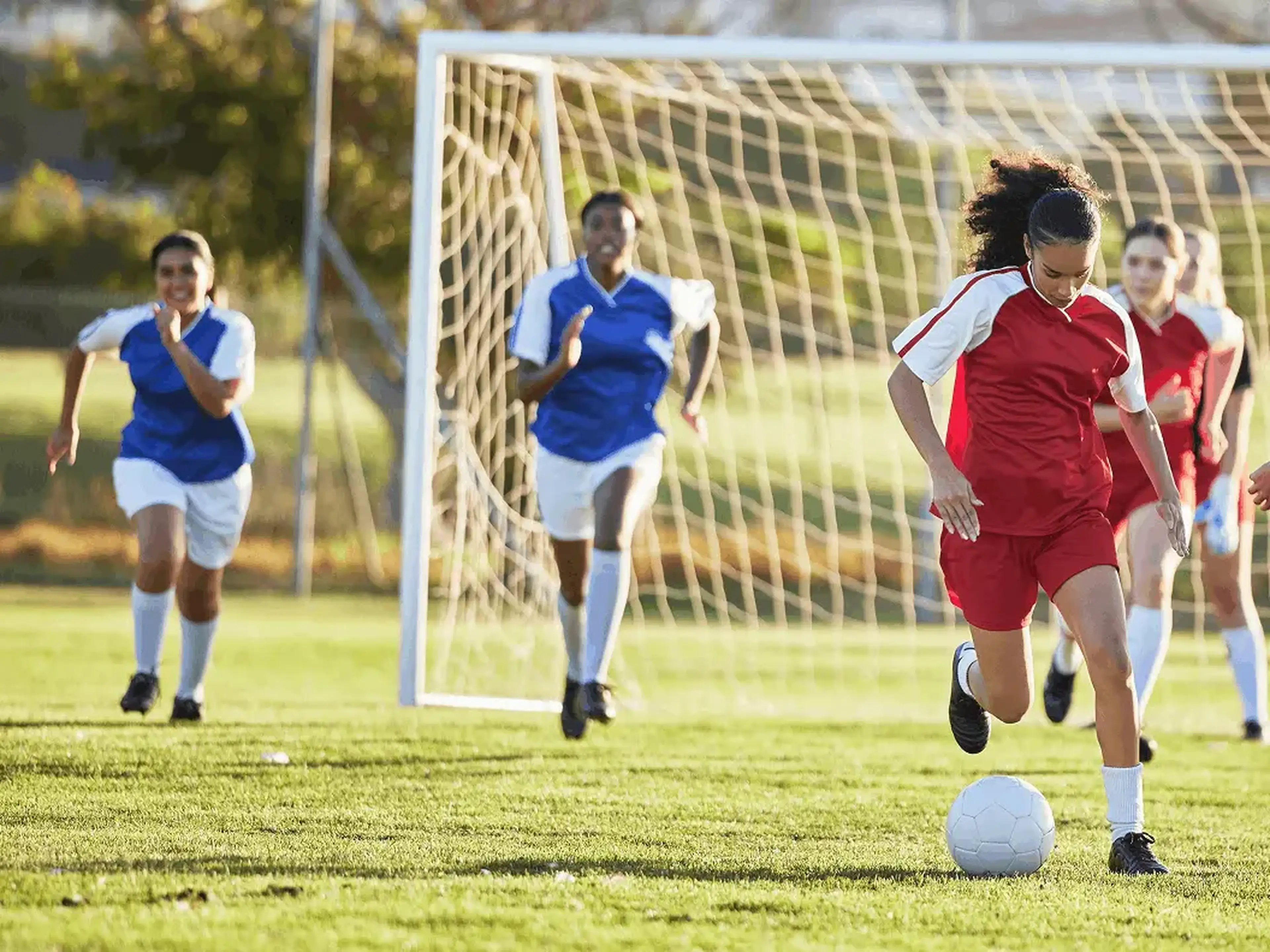 A soccer player in red dribbles a ball toward the goal, pursued by players in blue jerseys on a sunny field.