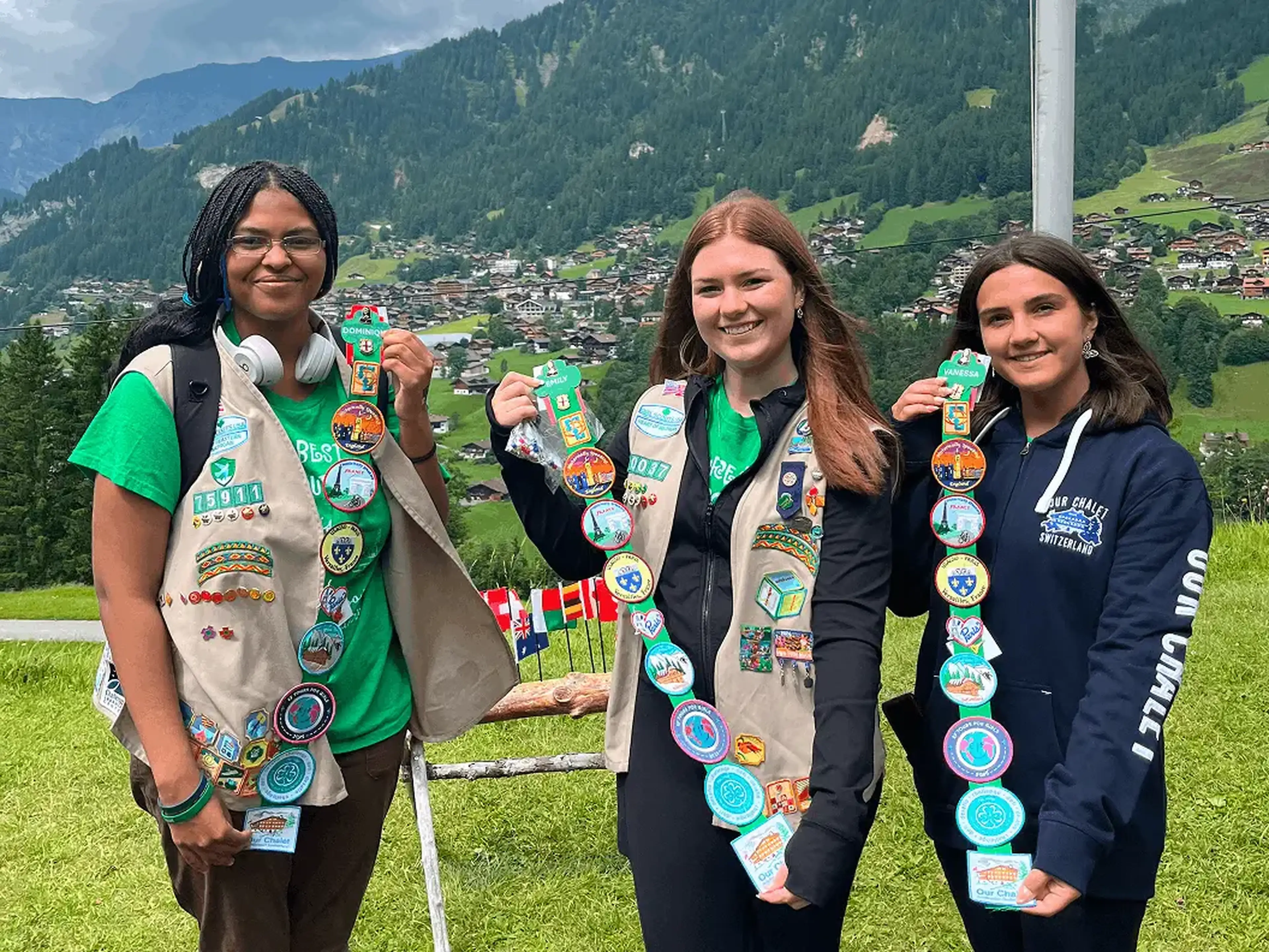 Three women outdoors hold sashes adorned with colorful patches, against a backdrop of mountains and a valley with buildings.