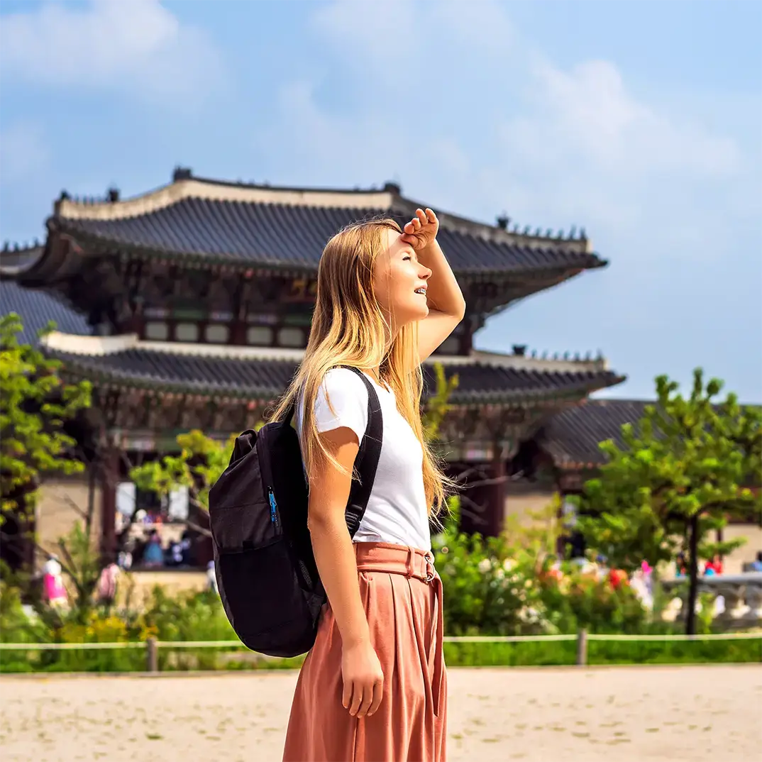 A student wearing a backpack shades her eyes while exploring a traditional palace in South Korea.