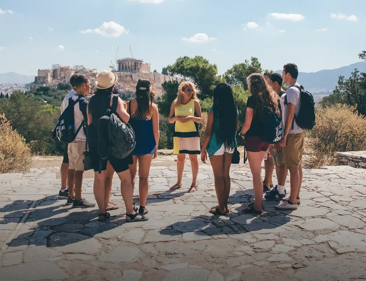 A Tour Director guide speaks to a group of students at an overlook with the Acropolis in the distance in Athens, Greece.