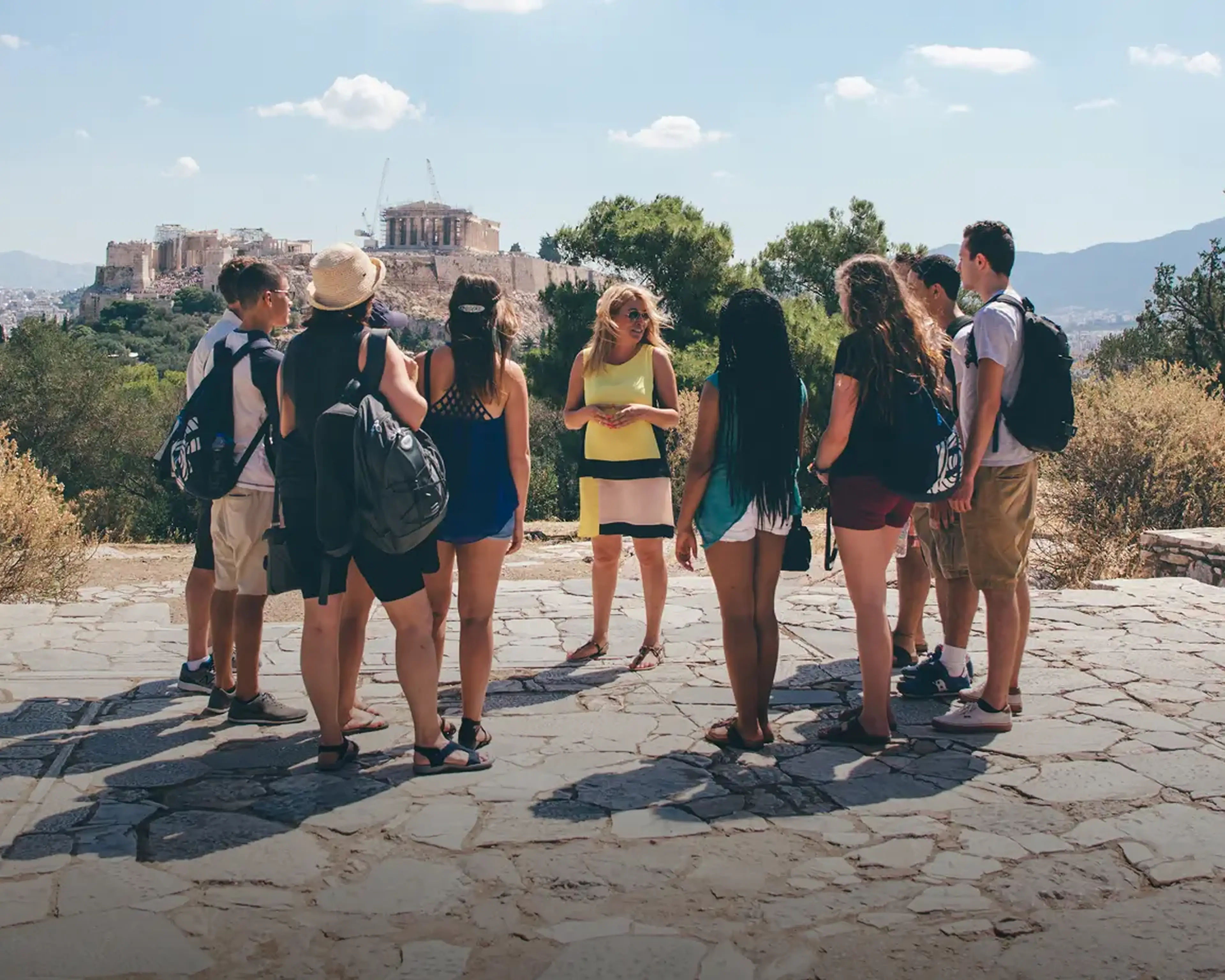 A Tour Director guide speaks to a group of students at an overlook with the Acropolis in the distance in Athens, Greece.