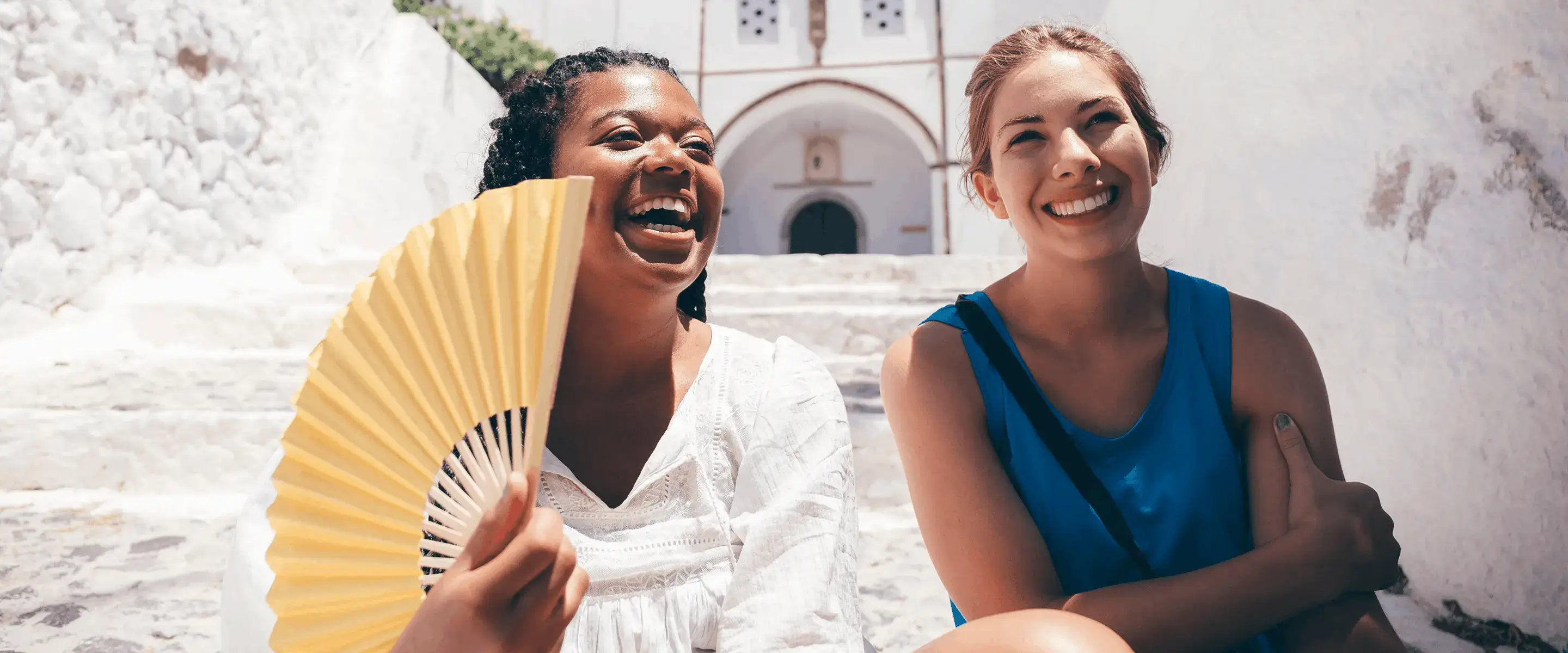 Two women sitting on stone steps, smiling; one holds a yellow fan. Bright, sunny setting with white buildings in the background.