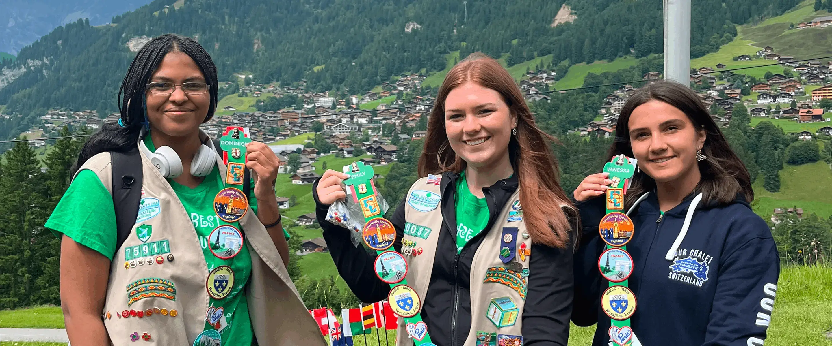 Three women in scout uniforms display embroidered patches, standing in front of a scenic mountain village. Various national flags are visible.