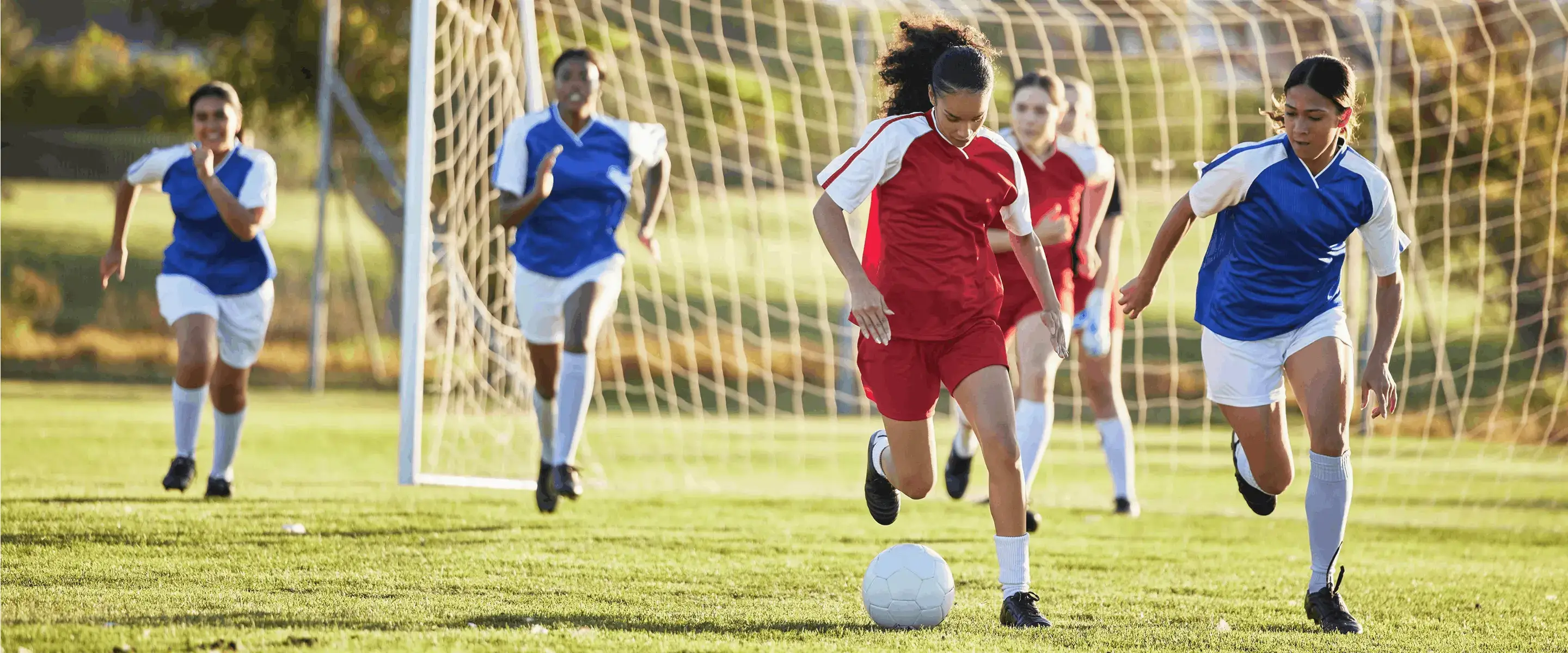 A soccer game with a player in red dribbling the ball, pursued by players in blue, near the goal in a sunny field.