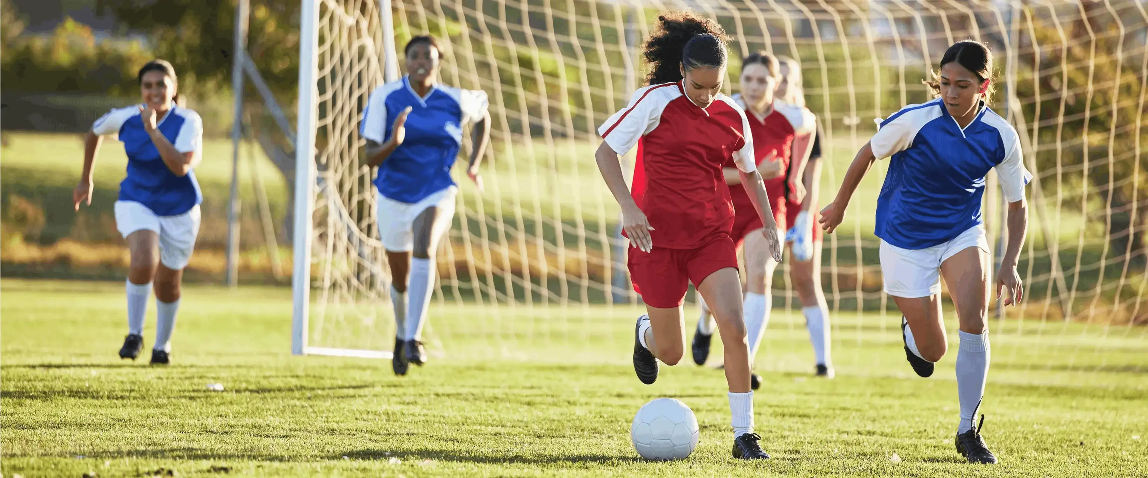 A soccer game with a player in red dribbling the ball, pursued by players in blue, near the goal in a sunny field.