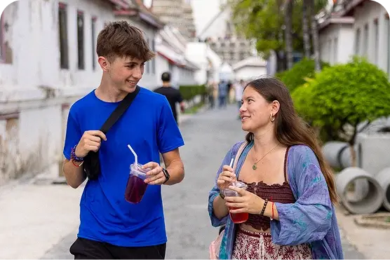 Two people walking outdoors, smiling at each other while holding drinks. A narrow street and greenery are in the background.
